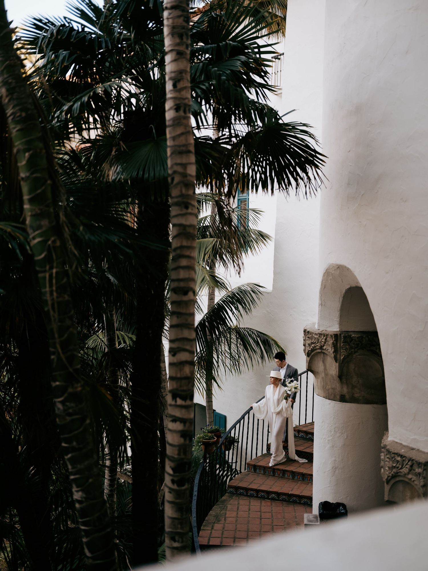 Wedding couple standing on spiral staircase surrounded by lush tropical palm trees and white walls.