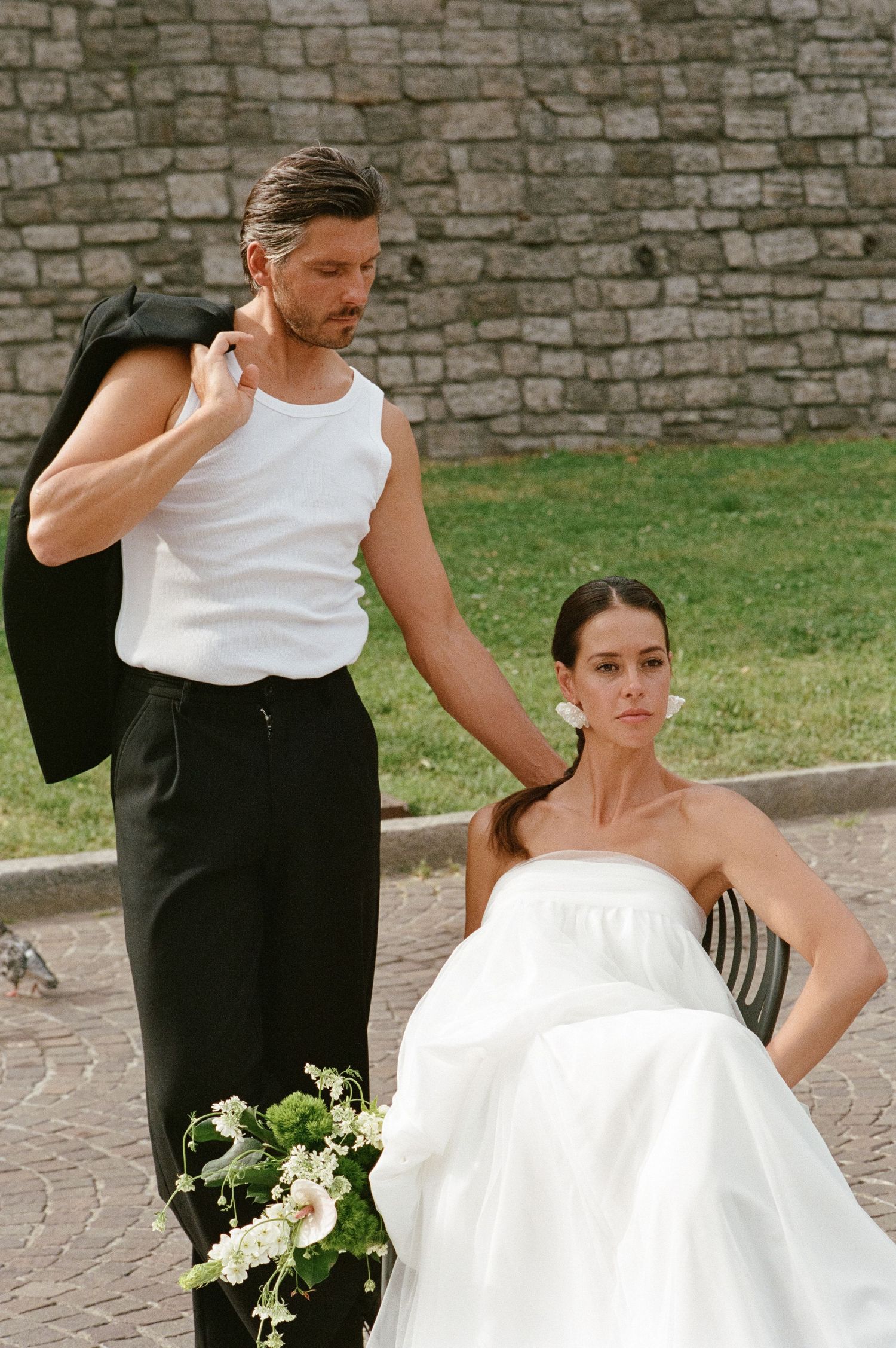 Groom in white tank top holds jacket over shoulder standing beside seated bride with green bouquet.