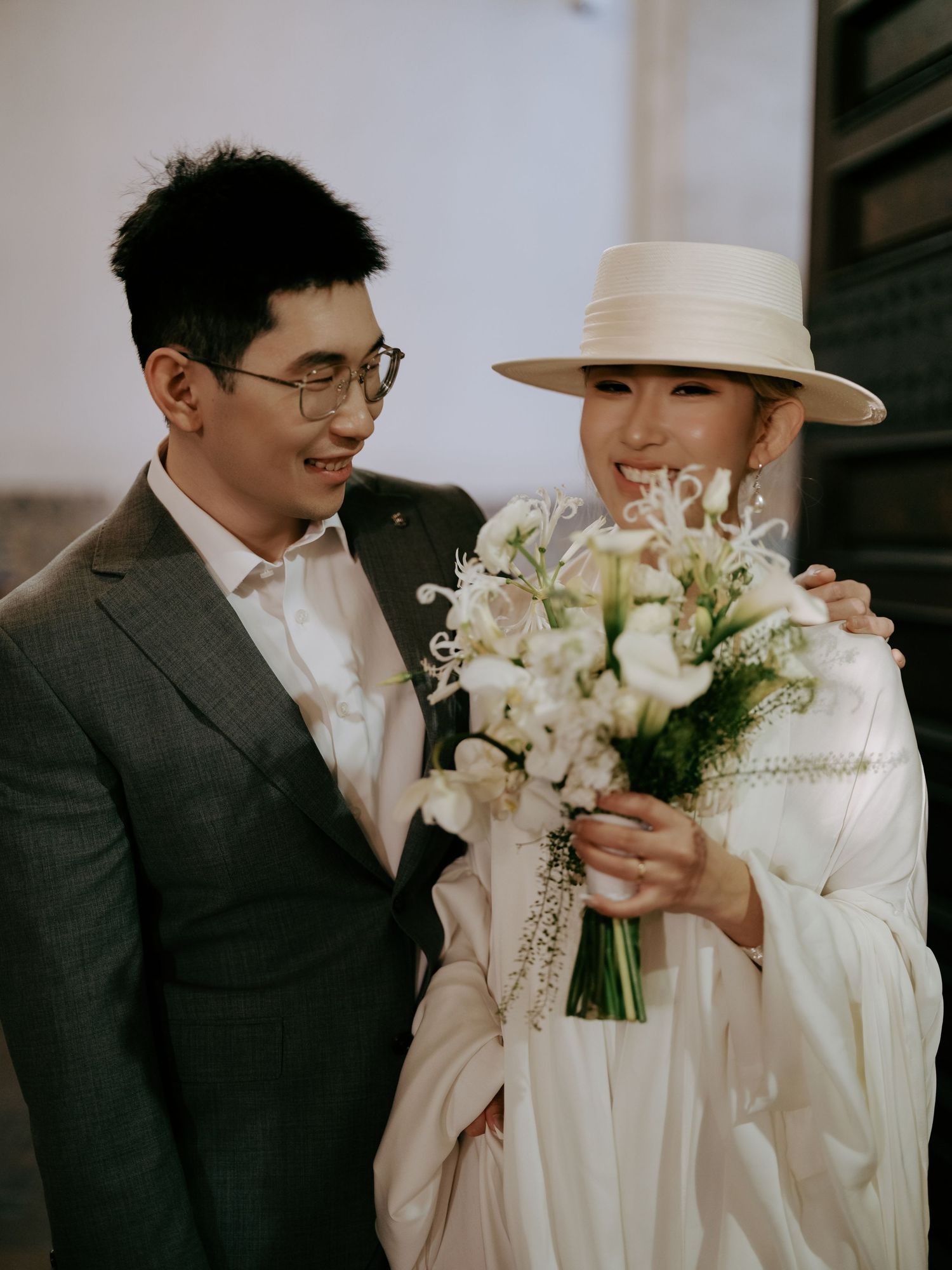 Smiling couple sharing a joyful moment, bride in white hat holding white floral wedding bouquet.
