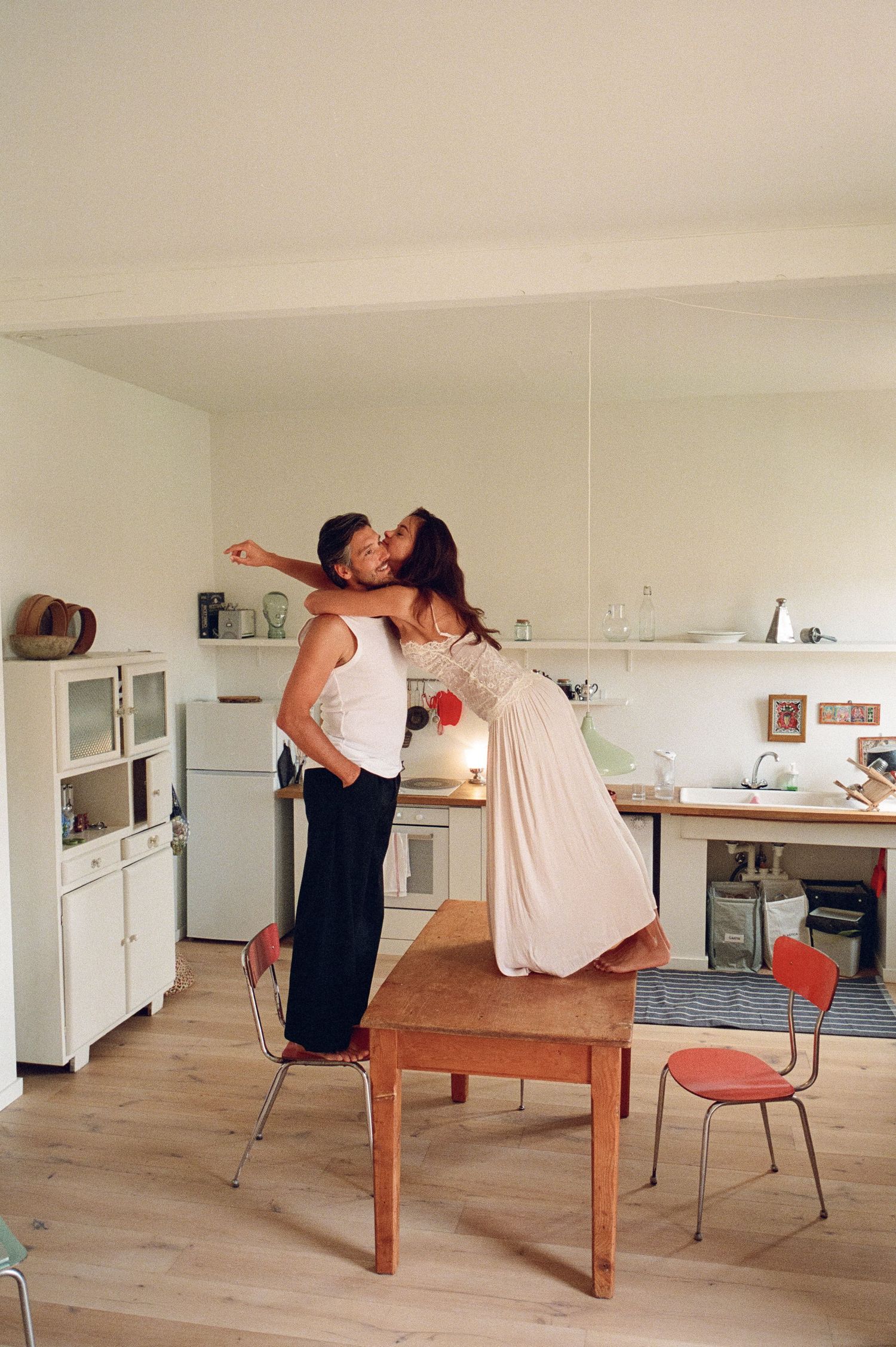 Couple sharing a tender kiss in a minimalist white kitchen, woman standing on wooden table.