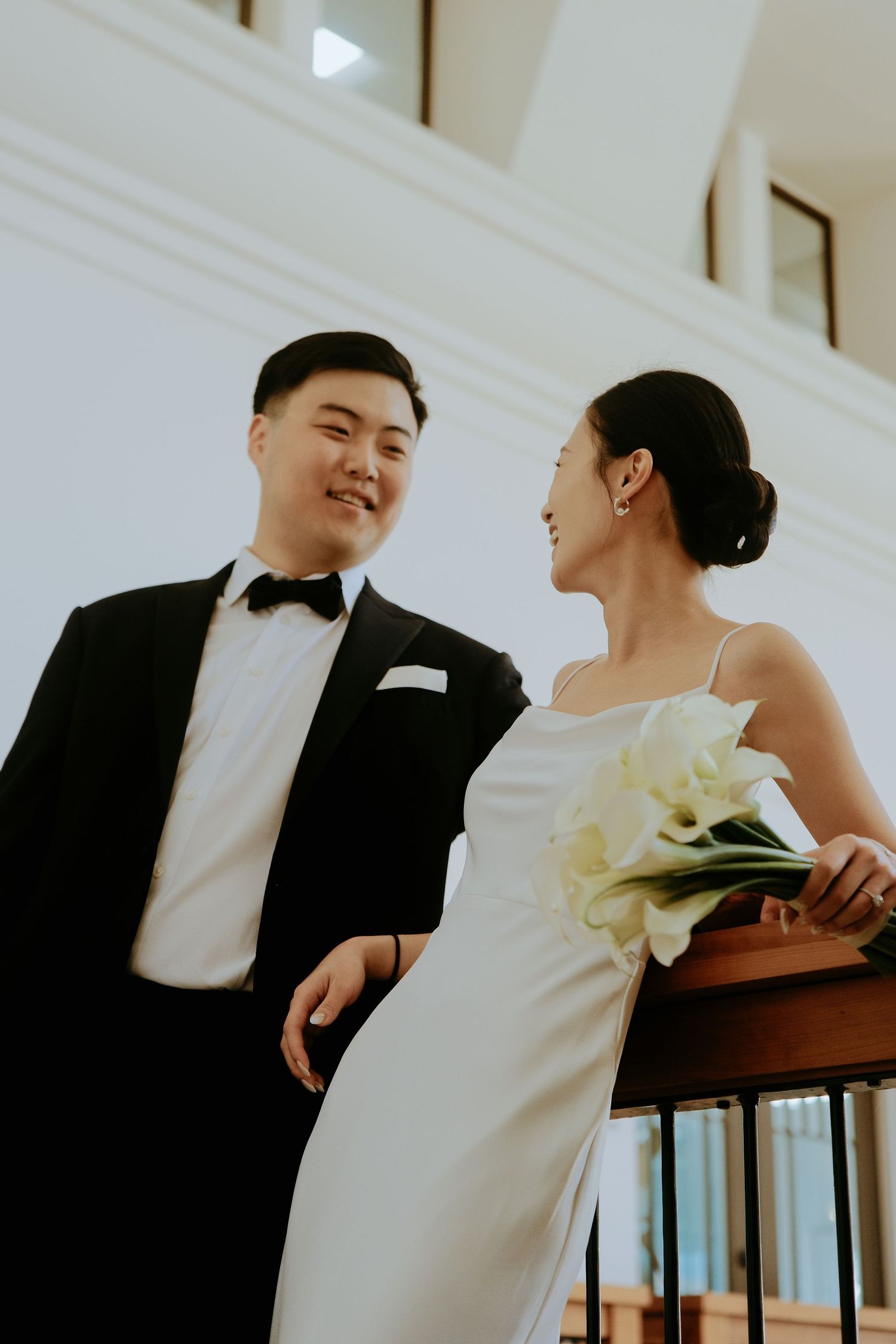 Asian bride and groom smiling at each other on balcony, bride in satin dress holding calla lilies, groom in tuxedo.