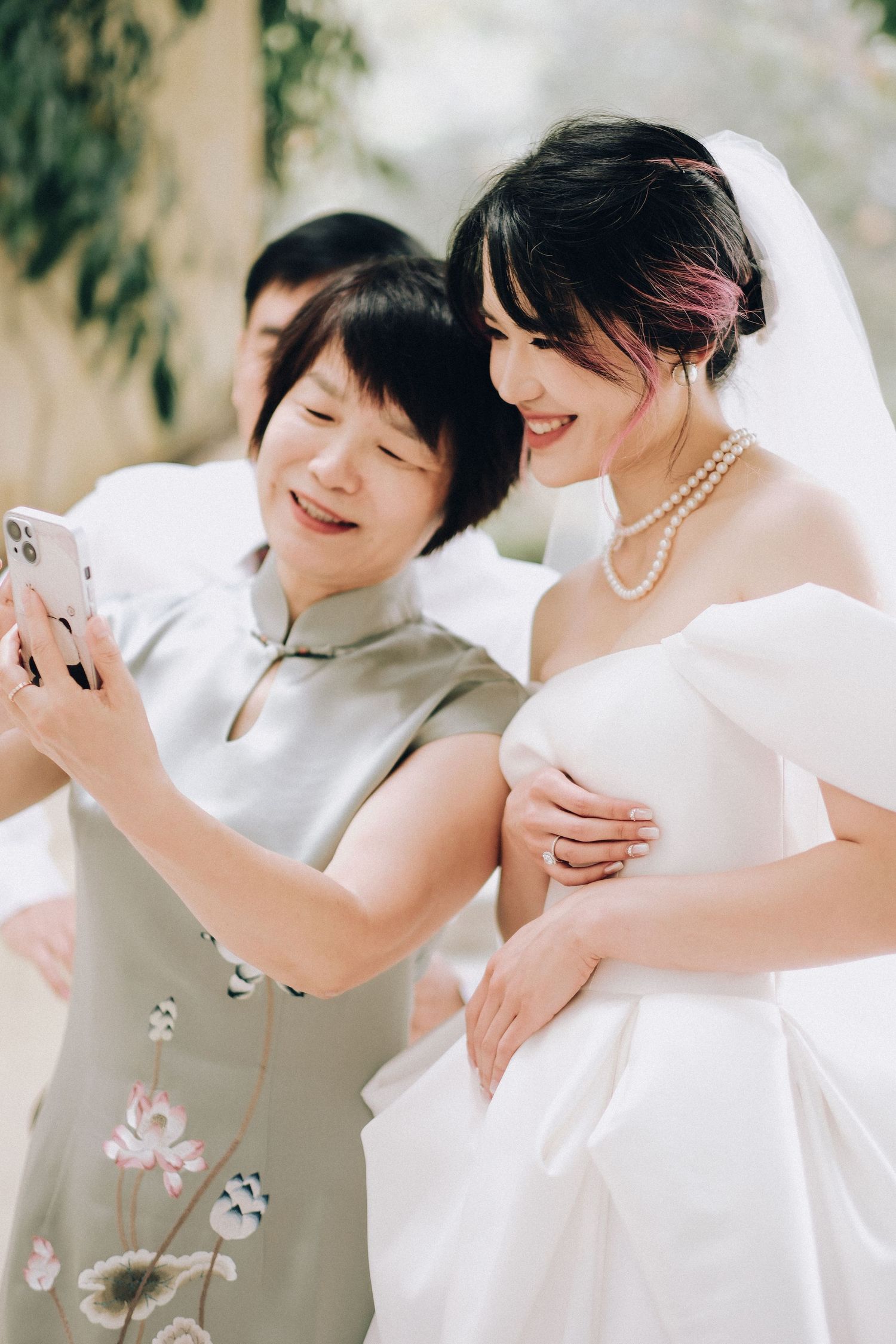 Bride in pearl necklace and white gown takes selfie with guest outdoors.