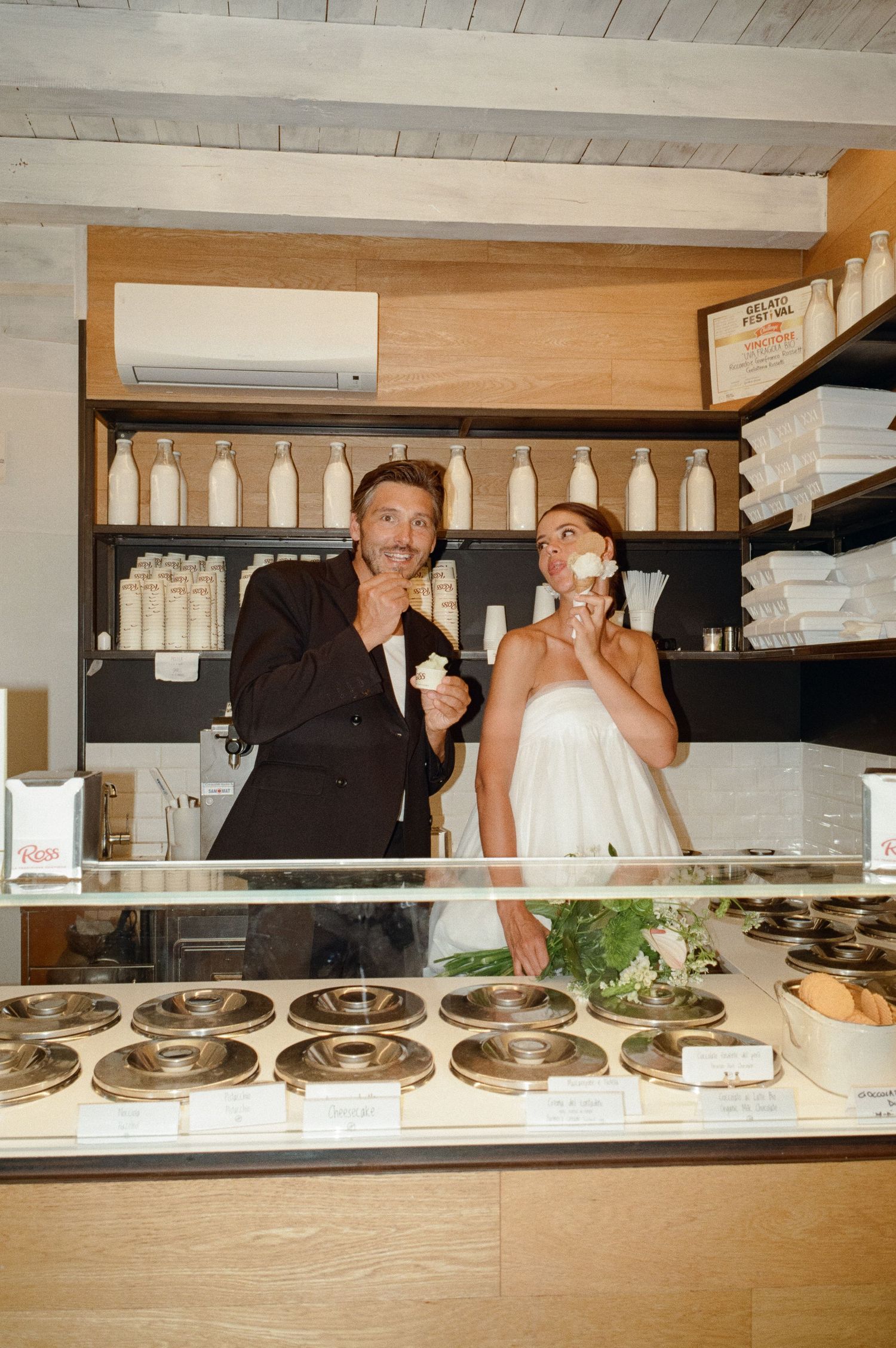 Bride and groom eating ice cream cones together behind a gelato shop display case in Italy.