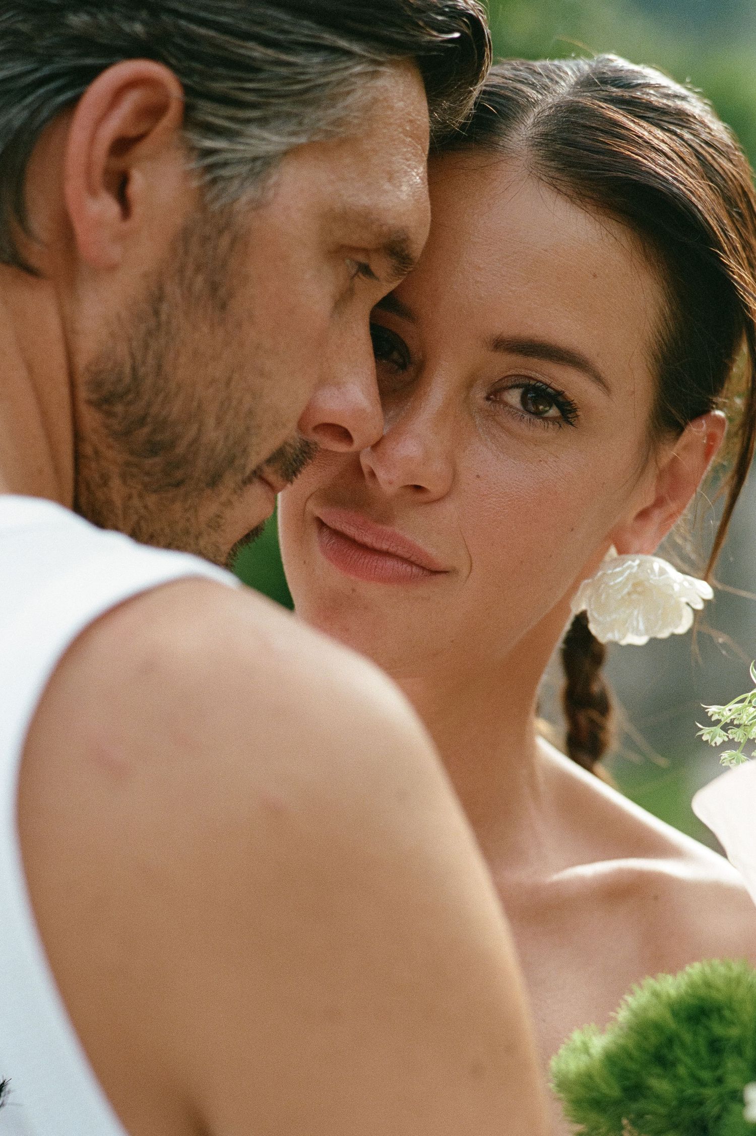 Bride with a flower earring and braid gazes softly as groom leans close in outdoor portrait.