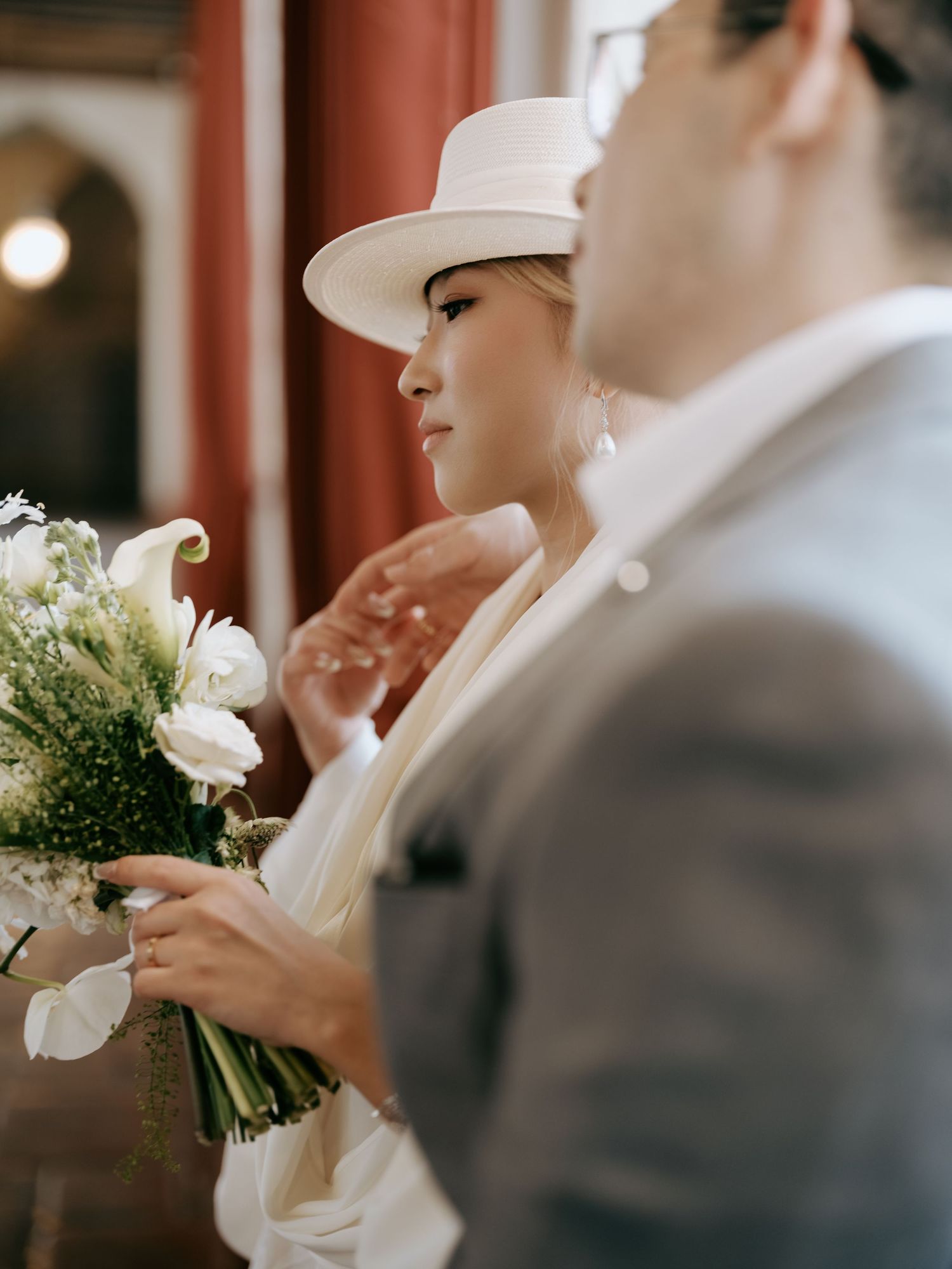 Close-up of bride in white hat holding white calla lily bouquet beside groom in gray suit.
