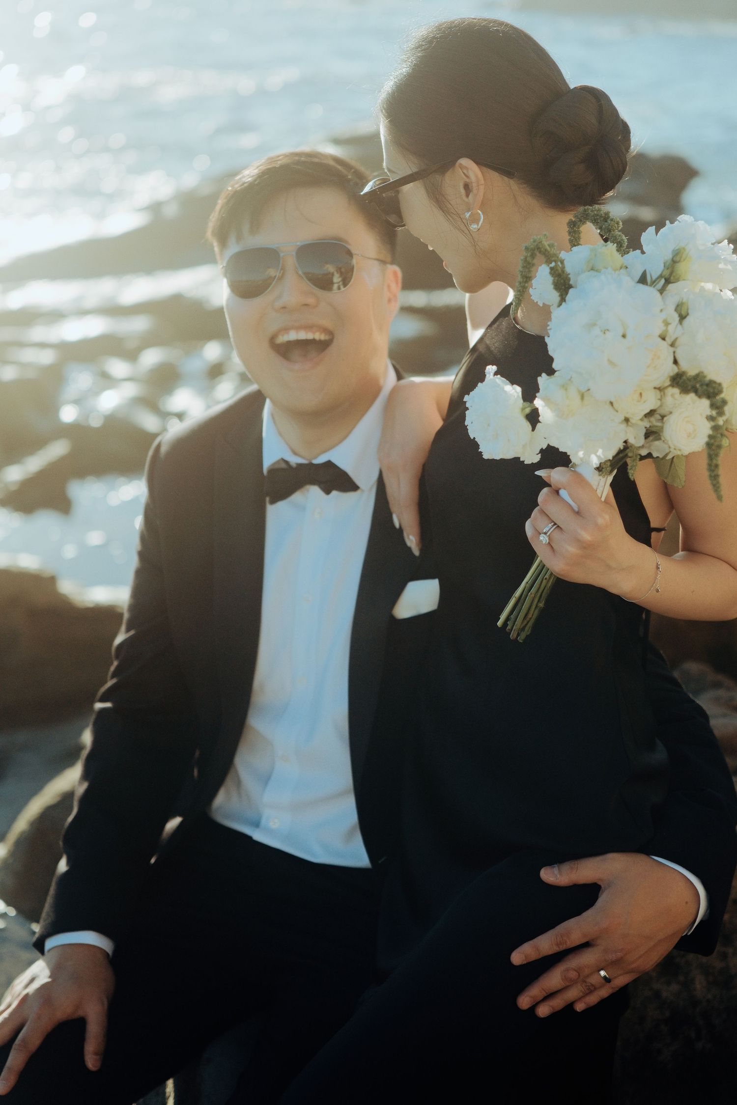 Laughing groom in tuxedo with bride holding white bouquet by ocean rocks.