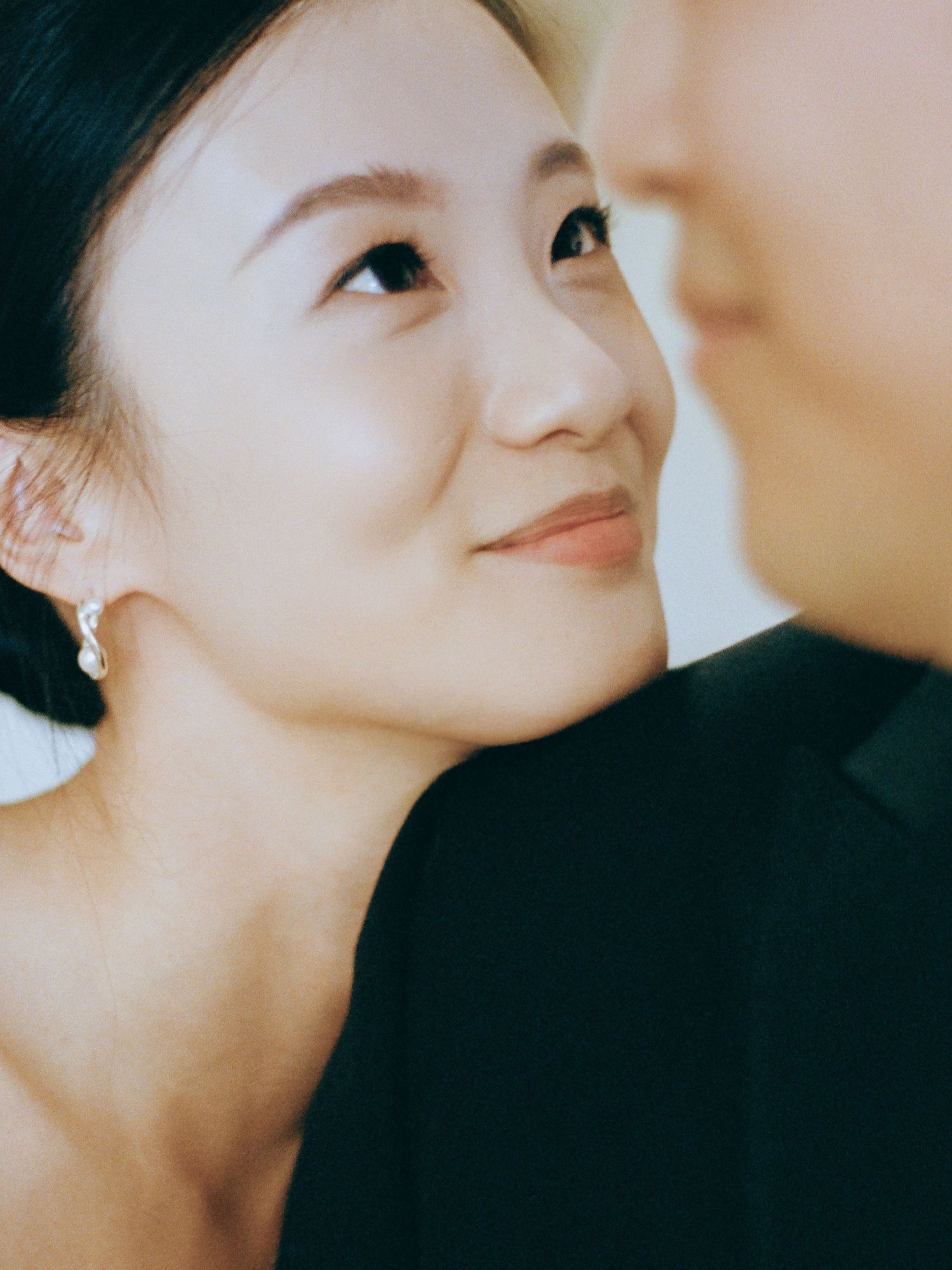 Close-up portrait of smiling Asian bride wearing pearl drop earrings with groom beside her.