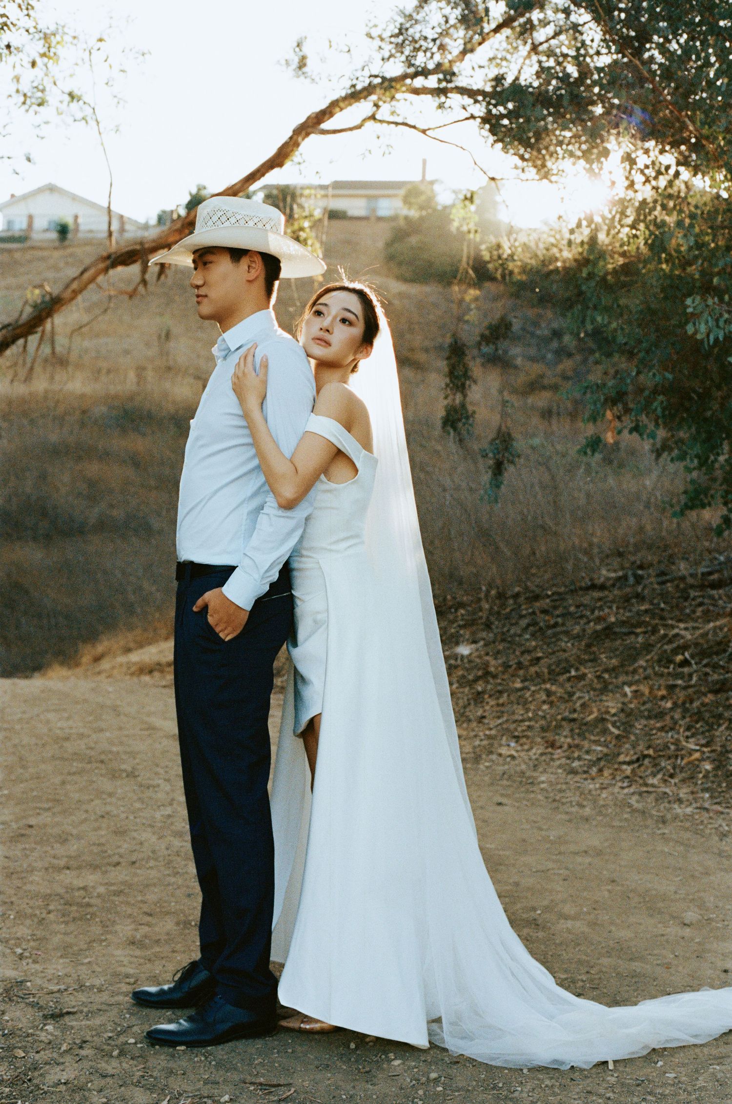 Bride in white gown and veil leans against groom in cowboy hat on a dry hillside path.