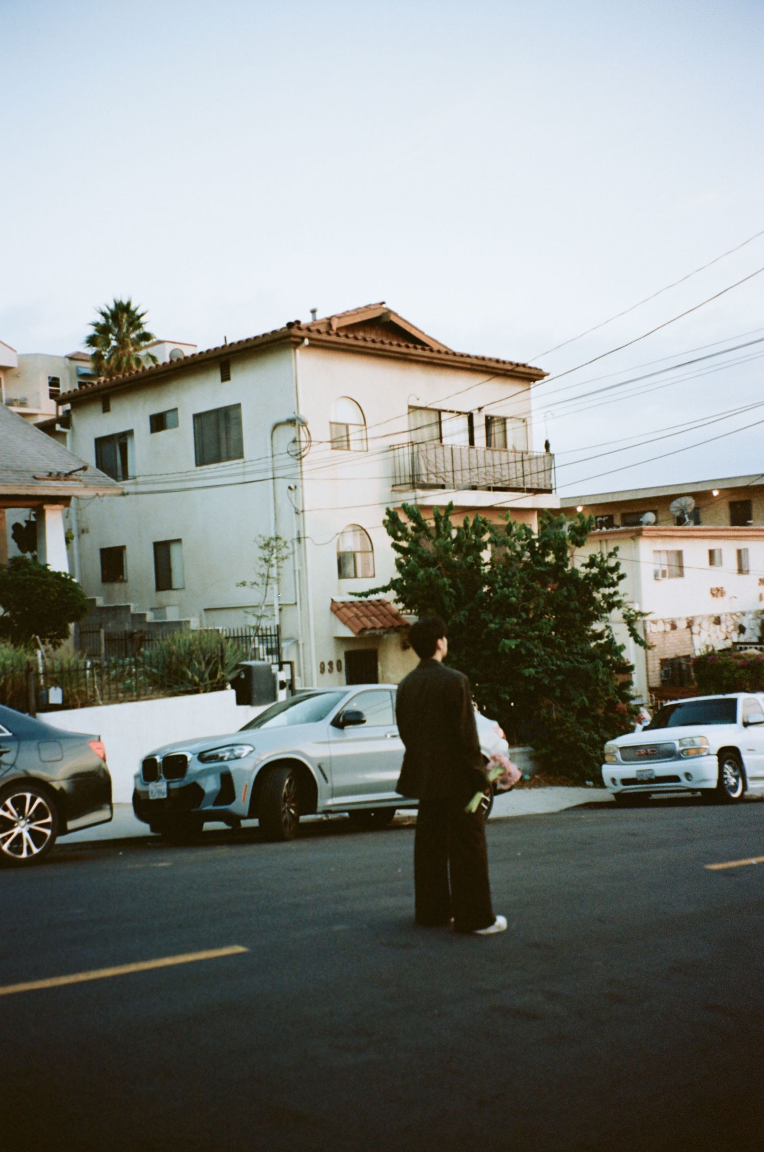 Groom in dark suit stands alone on suburban street with parked cars and Spanish-style buildings at dusk.