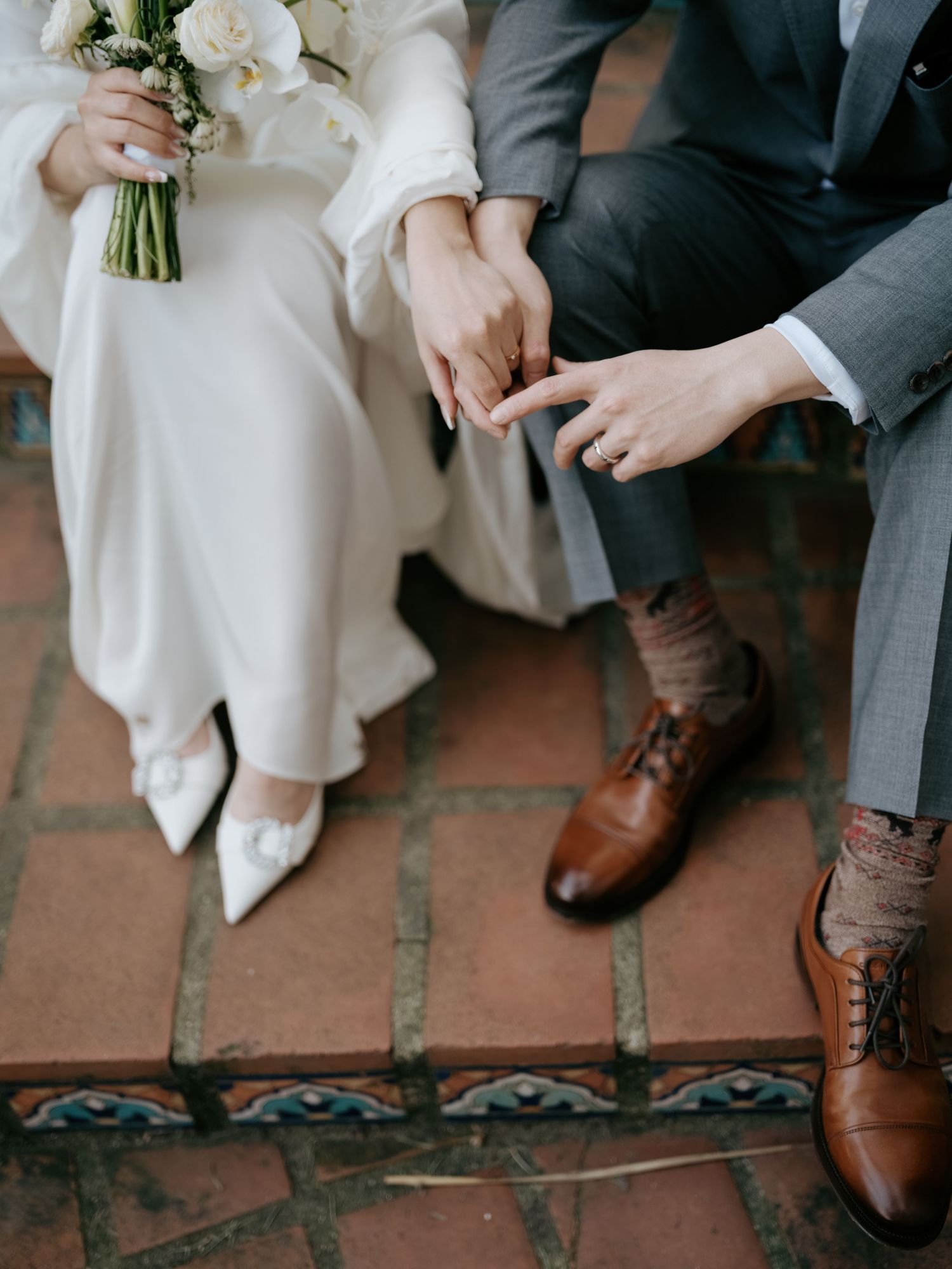 Close-up of bride and groom holding hands, showing white heels and brown oxford shoes.