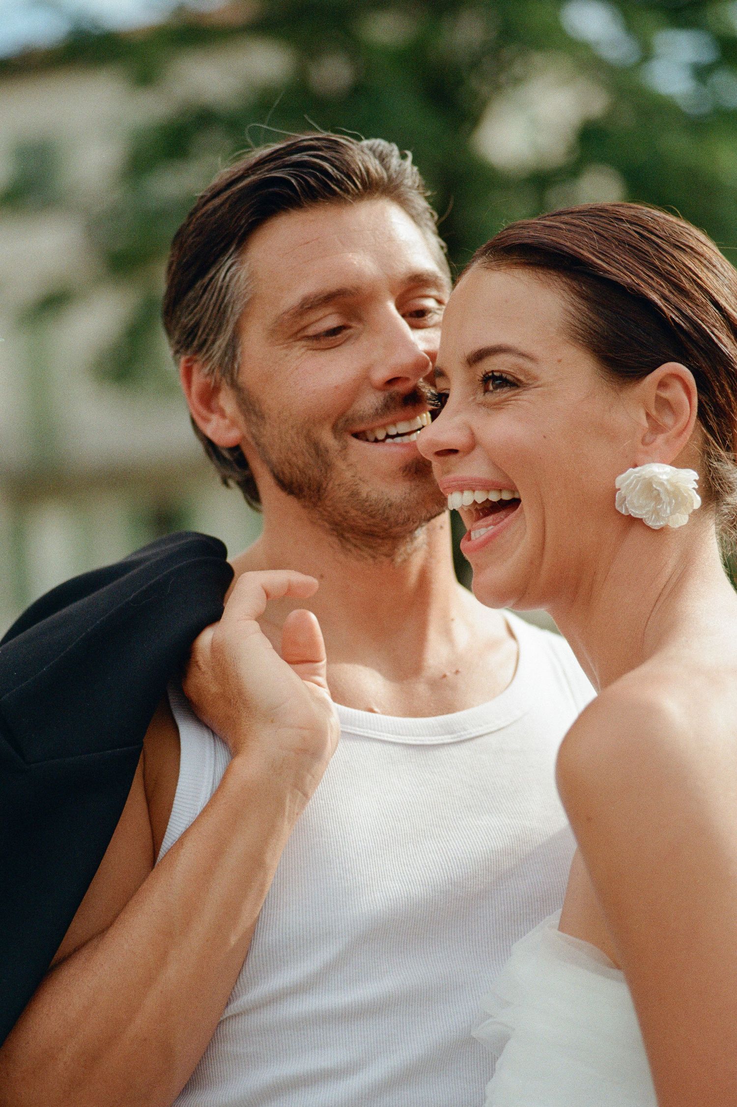 Laughing bride with floral earring and groom in white tank top sharing a joyful moment.