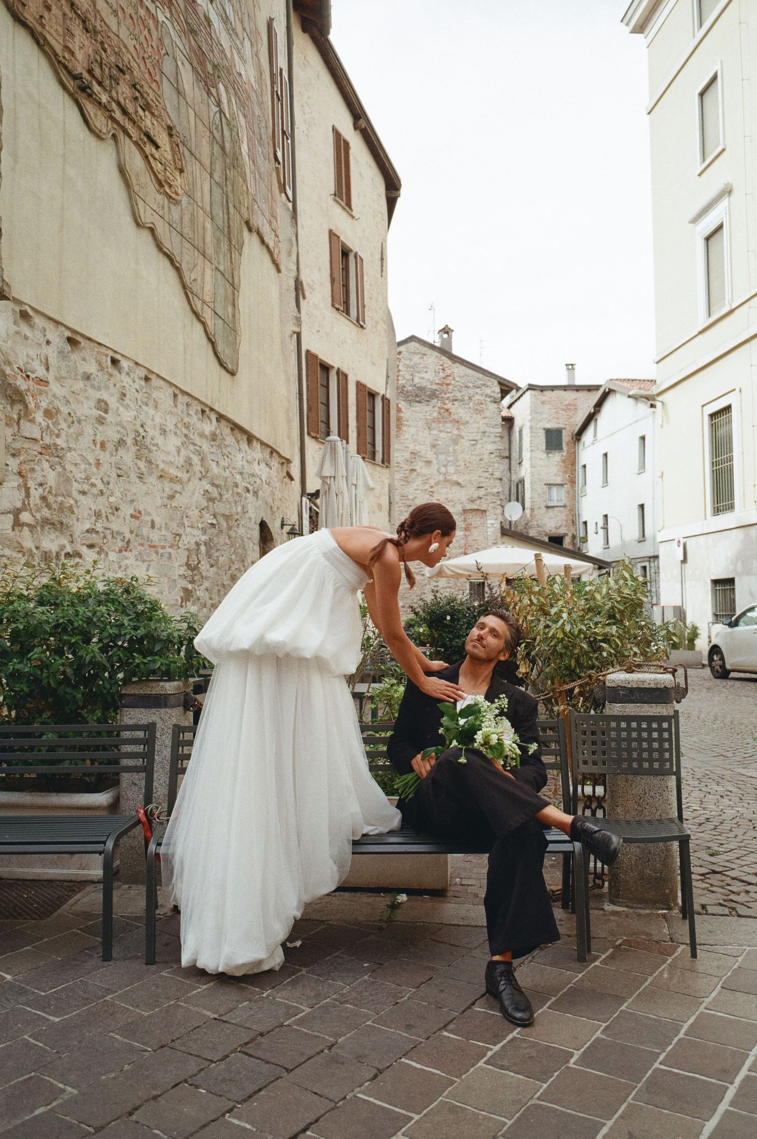 Bride leaning toward seated groom on a bench in a rustic Italian town square setting.