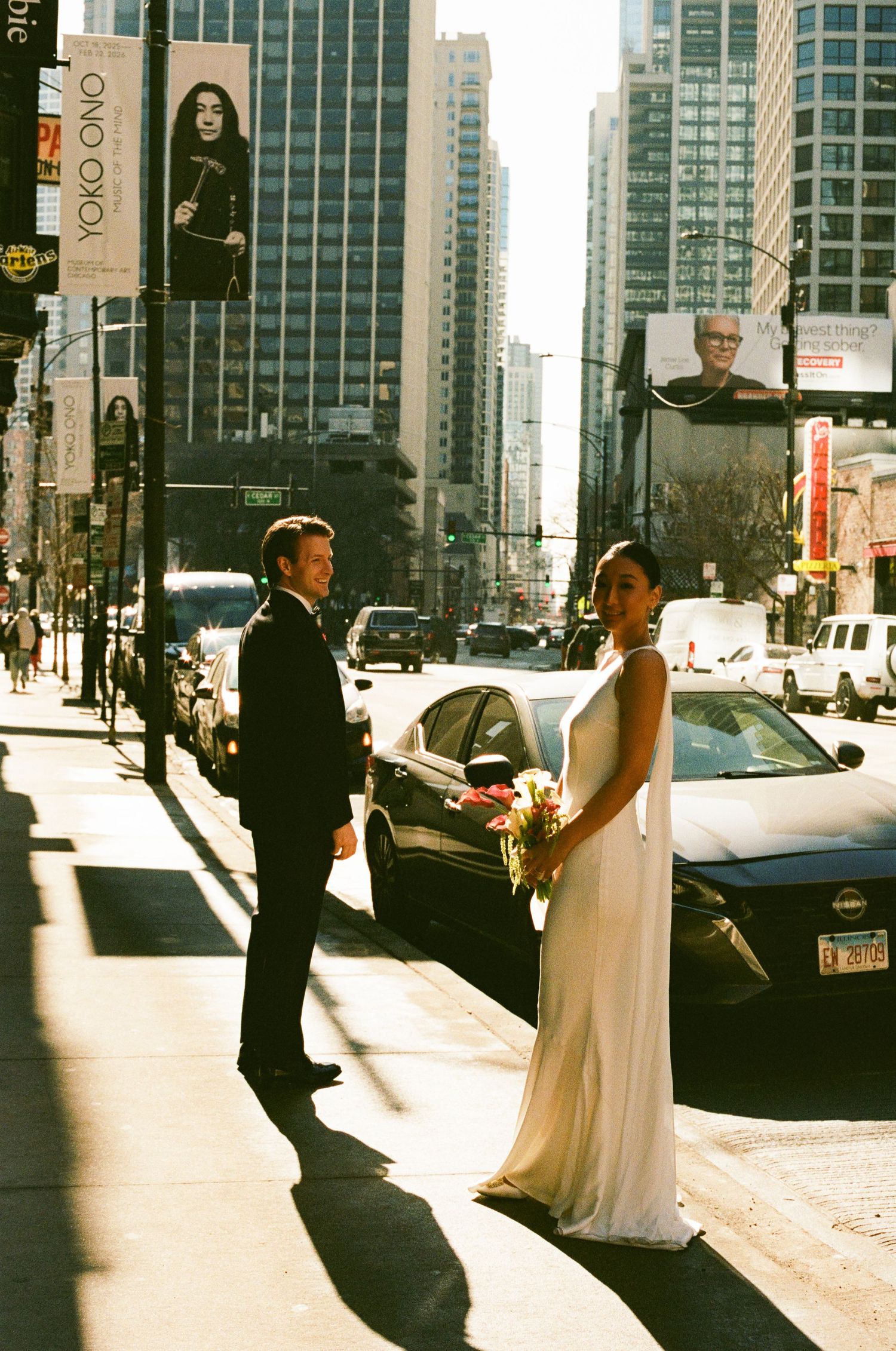 Bride in white gown and groom in tuxedo walk Chicago street with Yoko Ono billboard overhead.