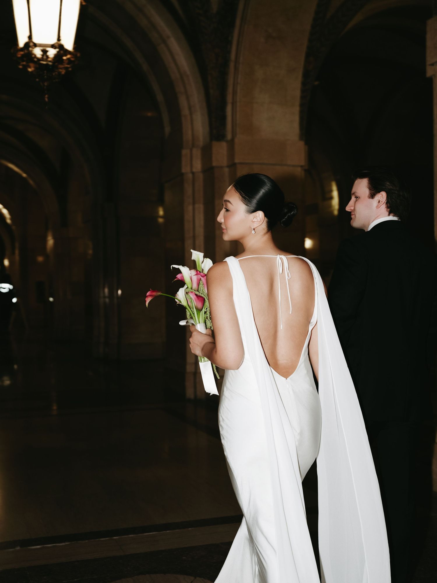 Elegant bride in backless white gown with cape holding pink bouquet in grand arched hallway.