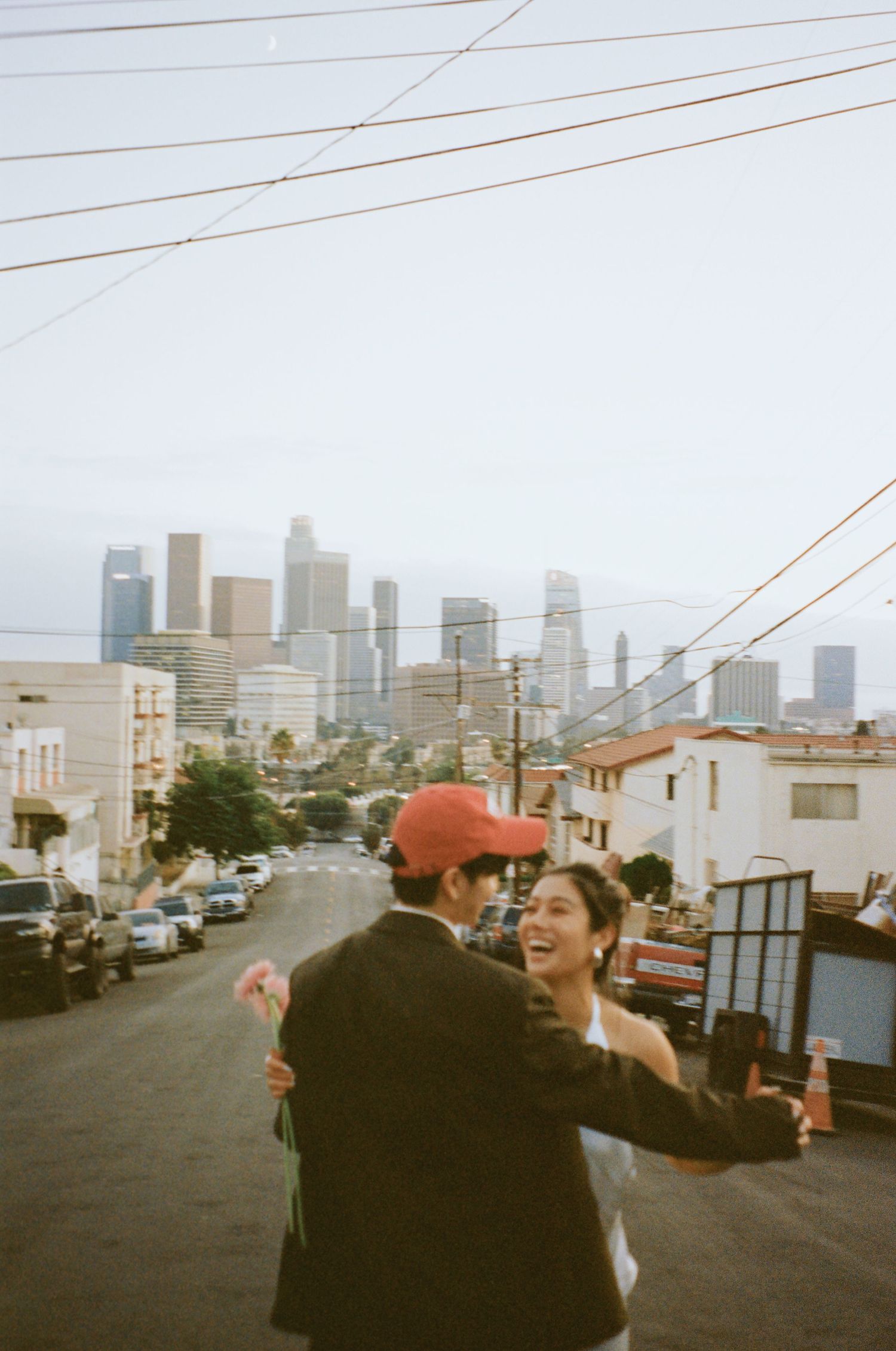 Laughing couple embracing on urban street hill with Los Angeles downtown skyline in the background.