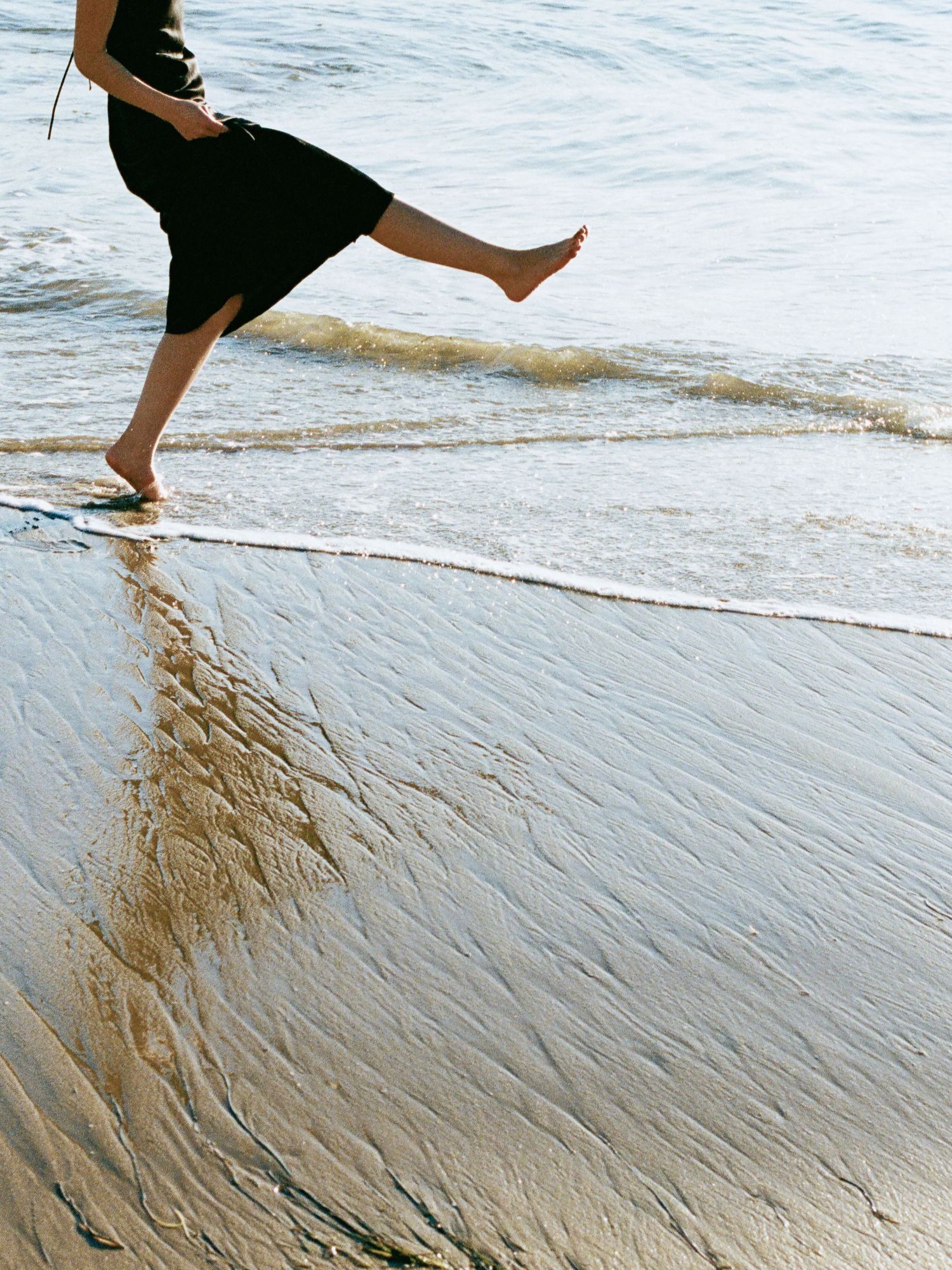 Woman in black dress kicking waves barefoot on sandy beach shoreline.