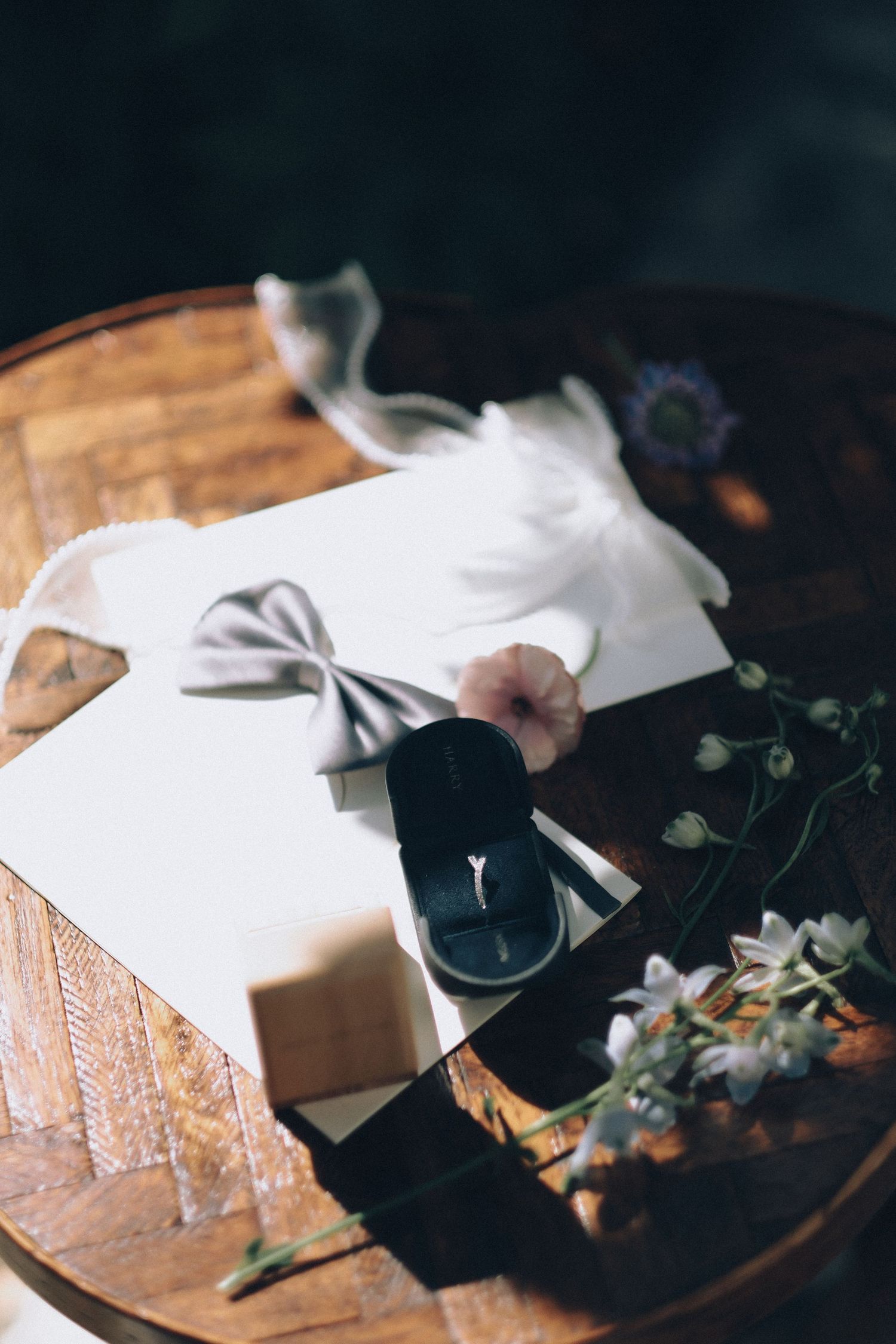 Wedding flat lay with engagement ring box, gray bow tie, invitation, and delicate flowers on wooden table.