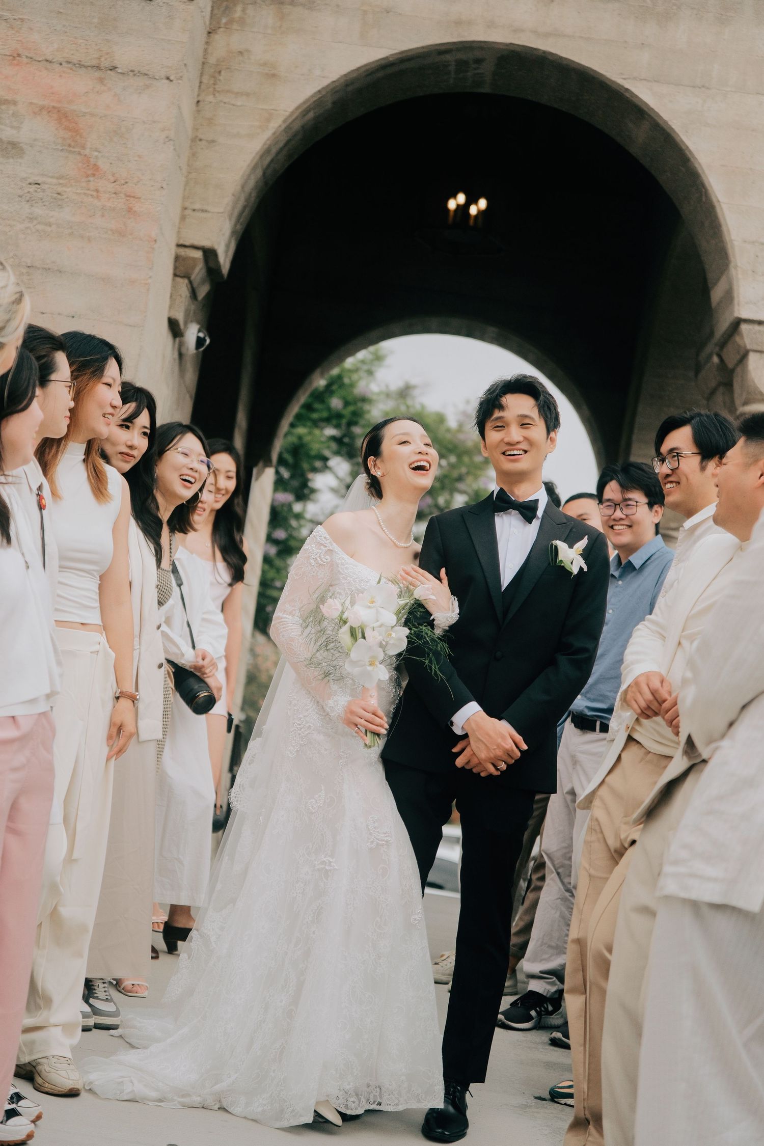 Joyful bride and groom exit ceremony arch surrounded by laughing wedding guests outdoors.