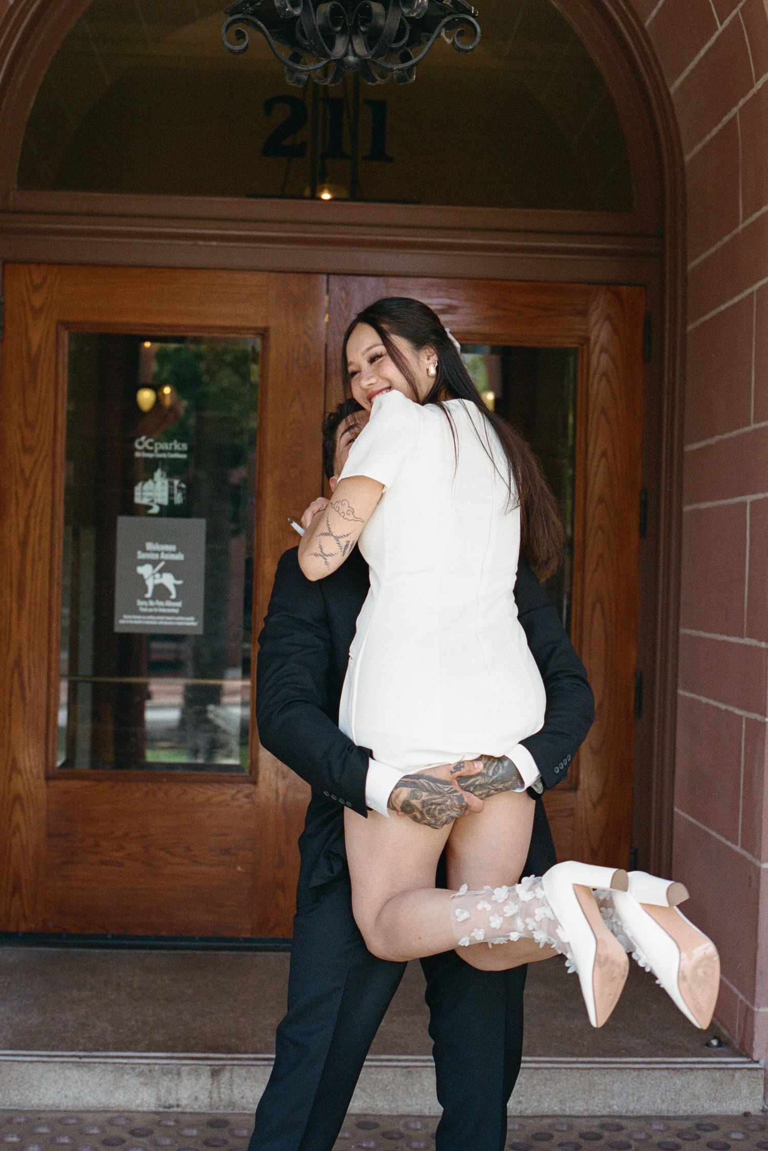 Groom in black suit lifts smiling bride in white mini dress outside a brick building entrance.