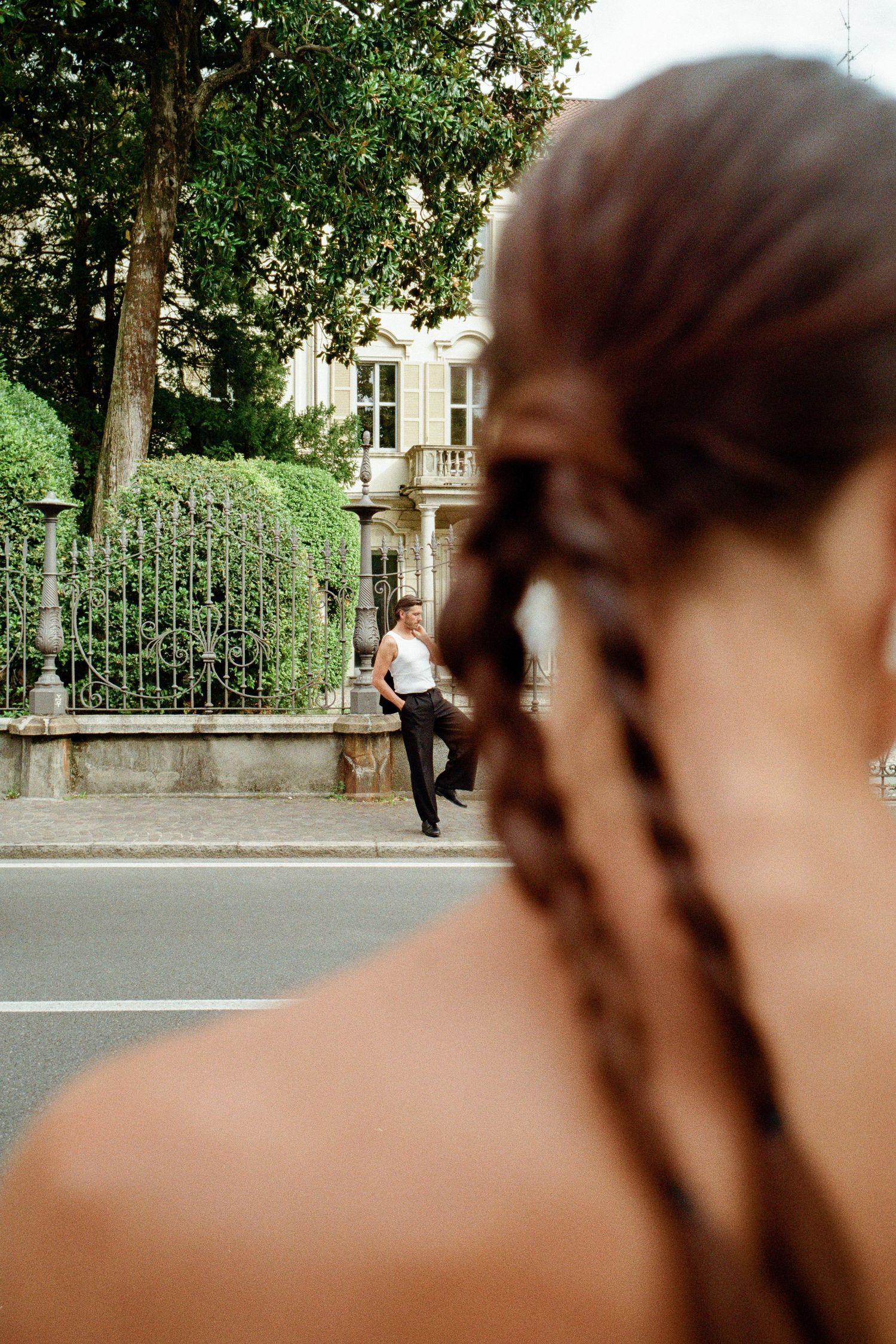 Bride with braided hair watching groom lean against ornate iron fence across the street.