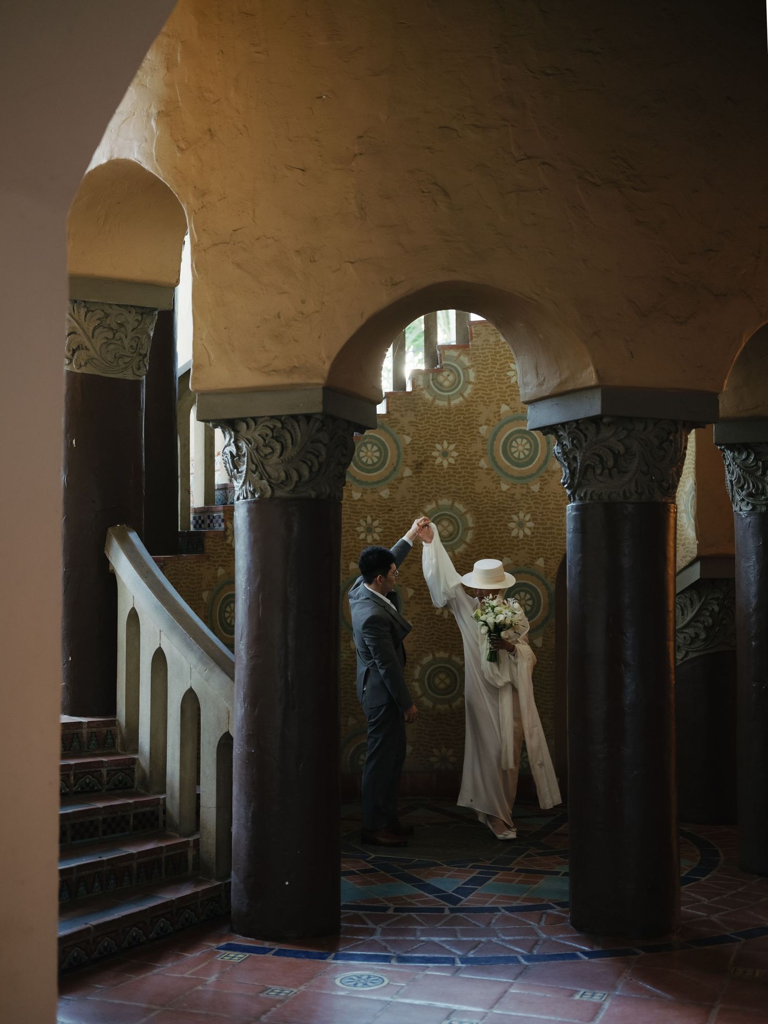 Groom twirls bride in white hat and coat under arched columns in elegant venue.