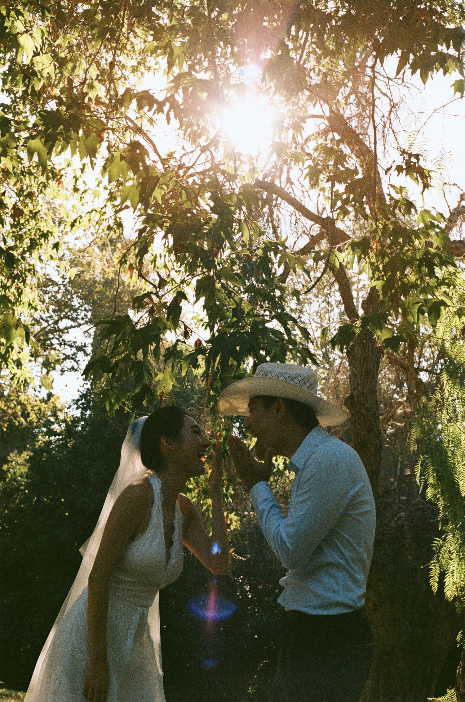 Bride and groom share a romantic moment under sunlit trees, groom in cowboy hat, with golden lens flare.