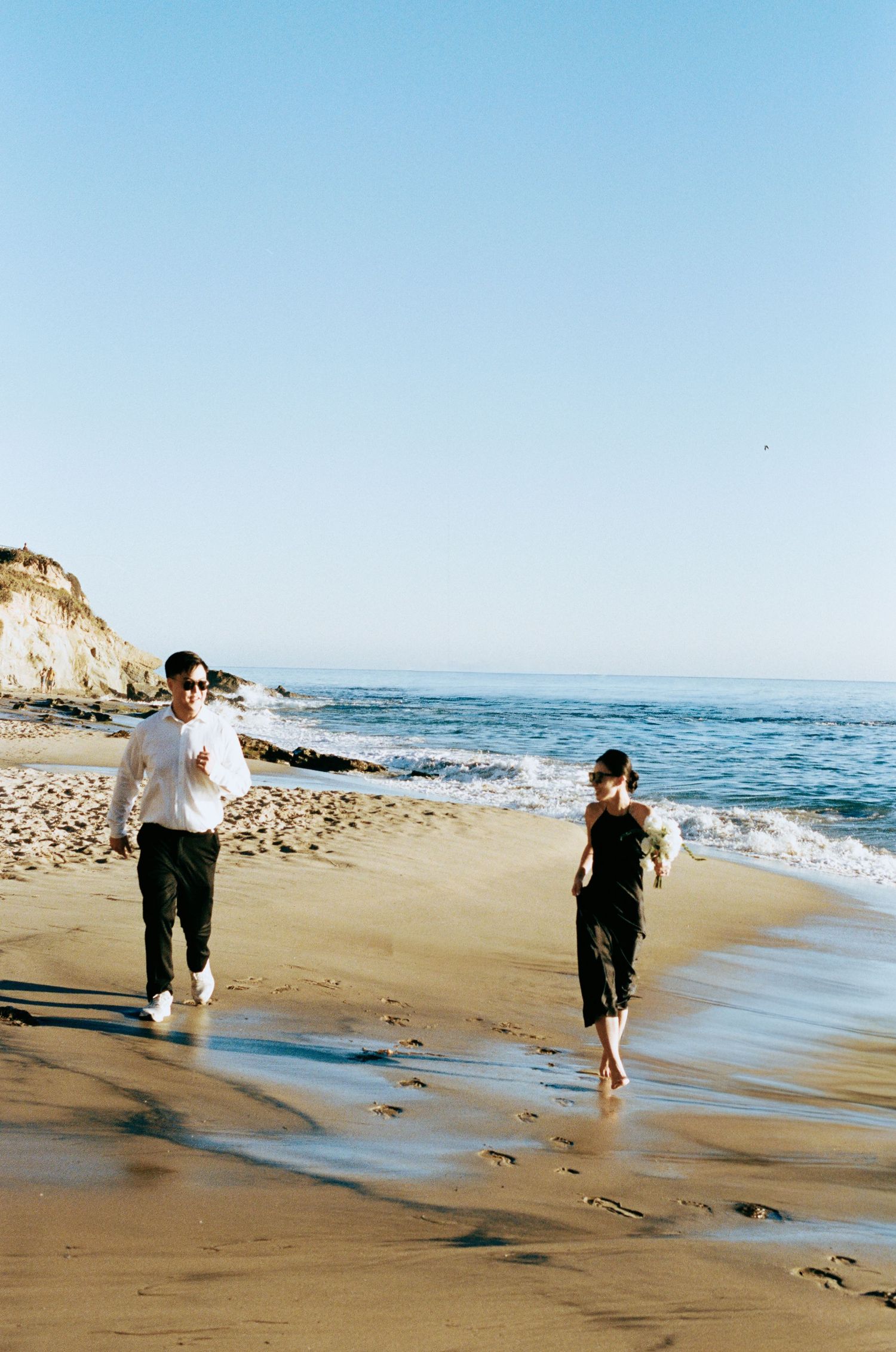 Bride and groom in casual attire walking along sandy beach shoreline on sunny day.