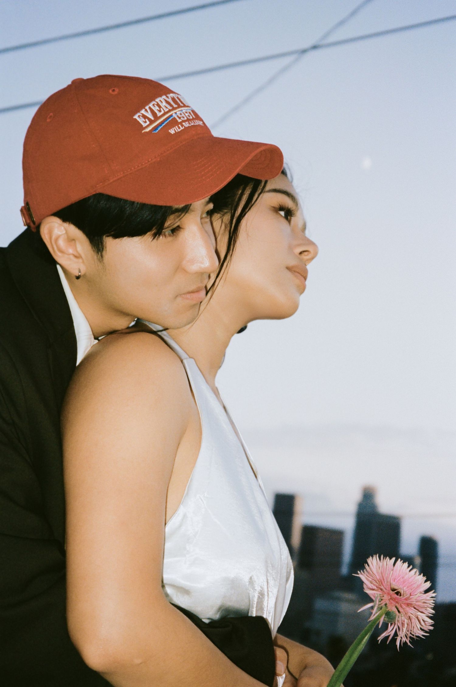 Couple embracing at golden hour, woman in white dress holding pink gerbera daisy, city skyline visible.