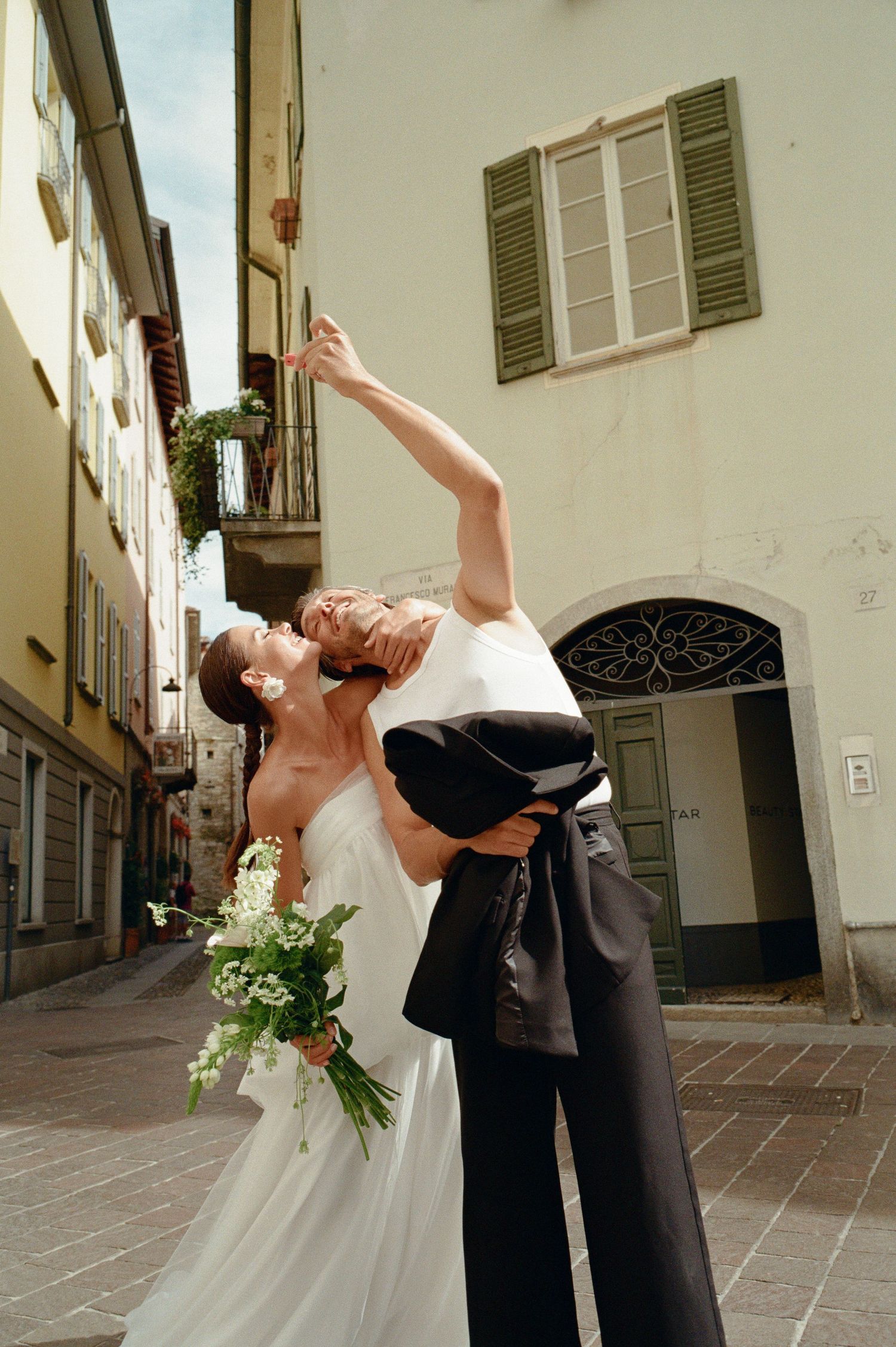 Groom dips bride holding bouquet of white flowers in a charming Italian cobblestone alley.