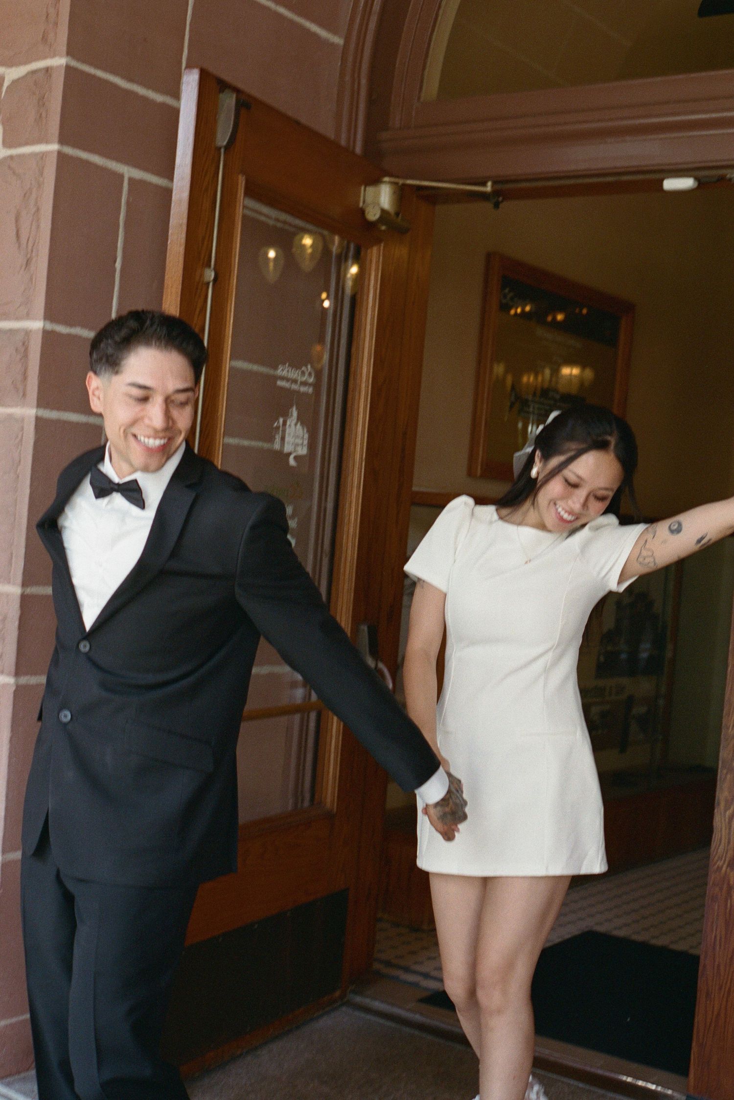 Laughing newlywed couple exits a brick building hand in hand after their wedding ceremony.