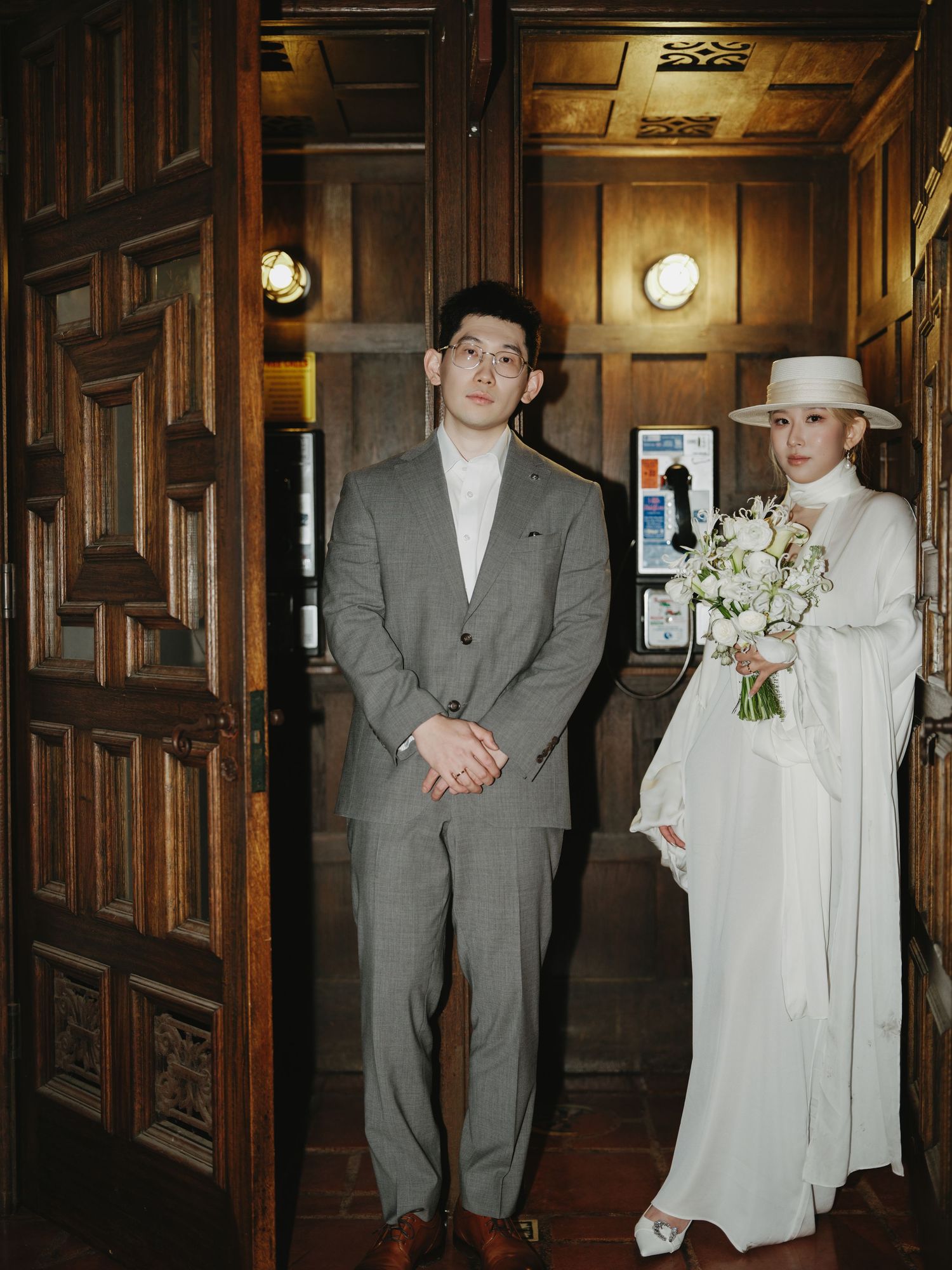 Bride in white coat and hat holds bouquet beside groom in gray suit at ornate wooden doors.