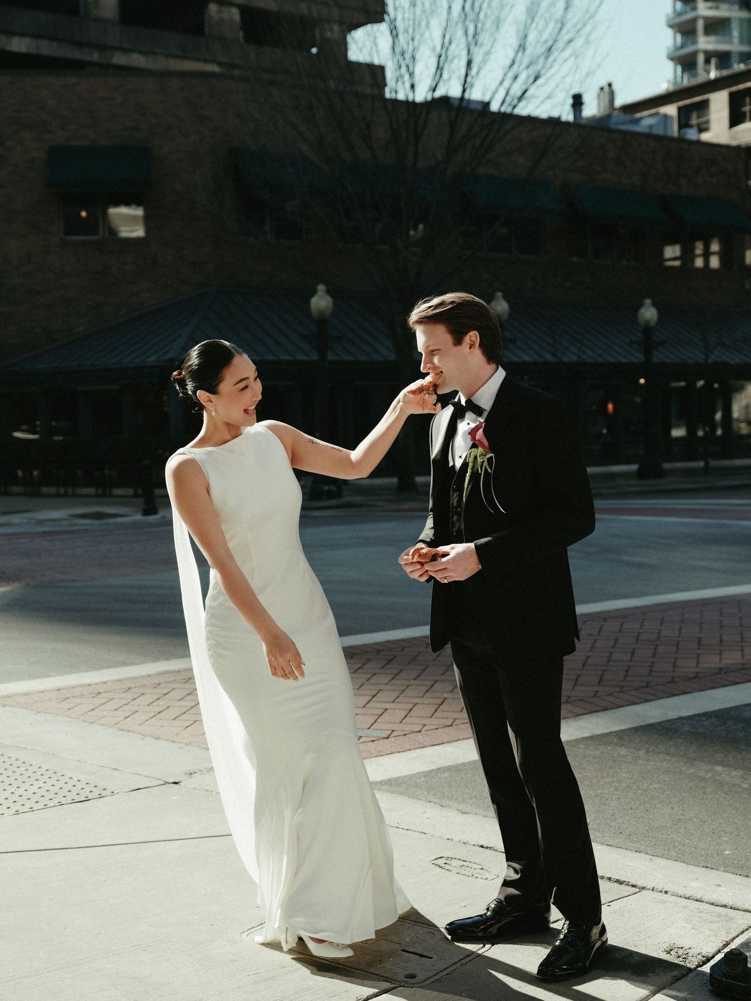 Bride playfully touches grooms face as they laugh together on a sunny urban street.