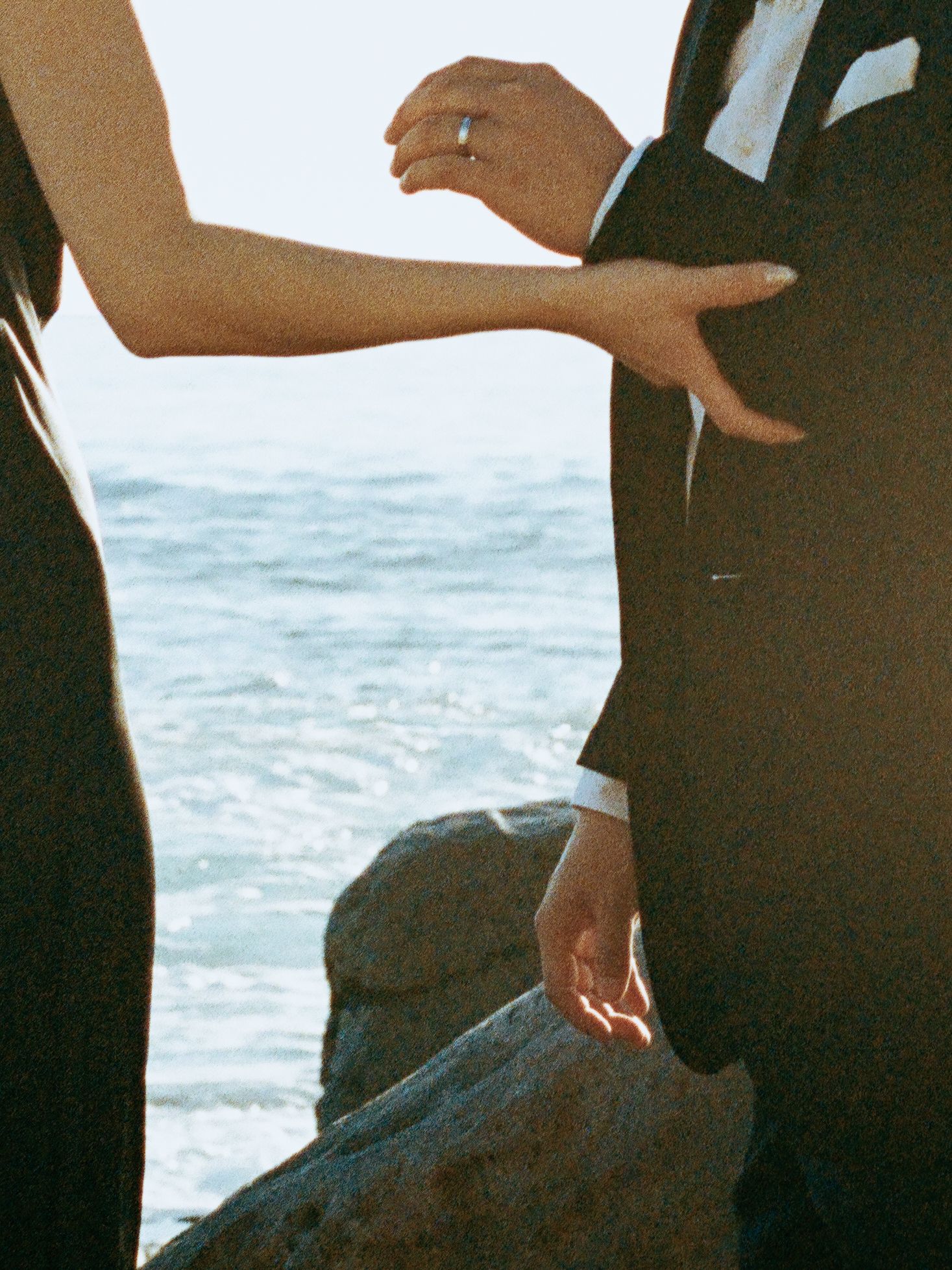Couple in formal black attire standing on coastal rocks with ocean waves in background.
