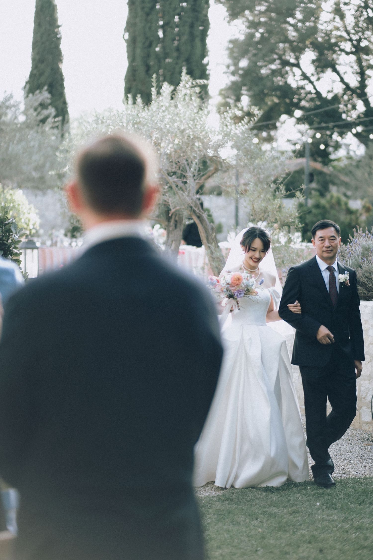 Bride walks down outdoor aisle with escort toward groom at garden wedding ceremony.