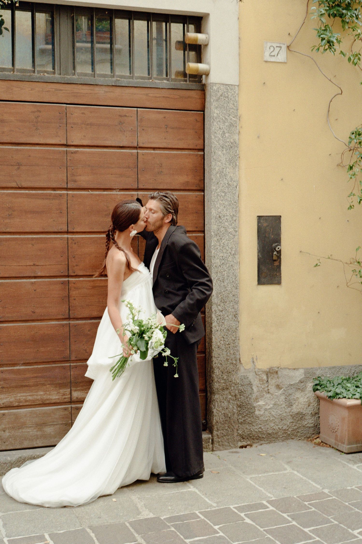 Bride and groom share a kiss in front of wooden garage door on Italian cobblestone street.