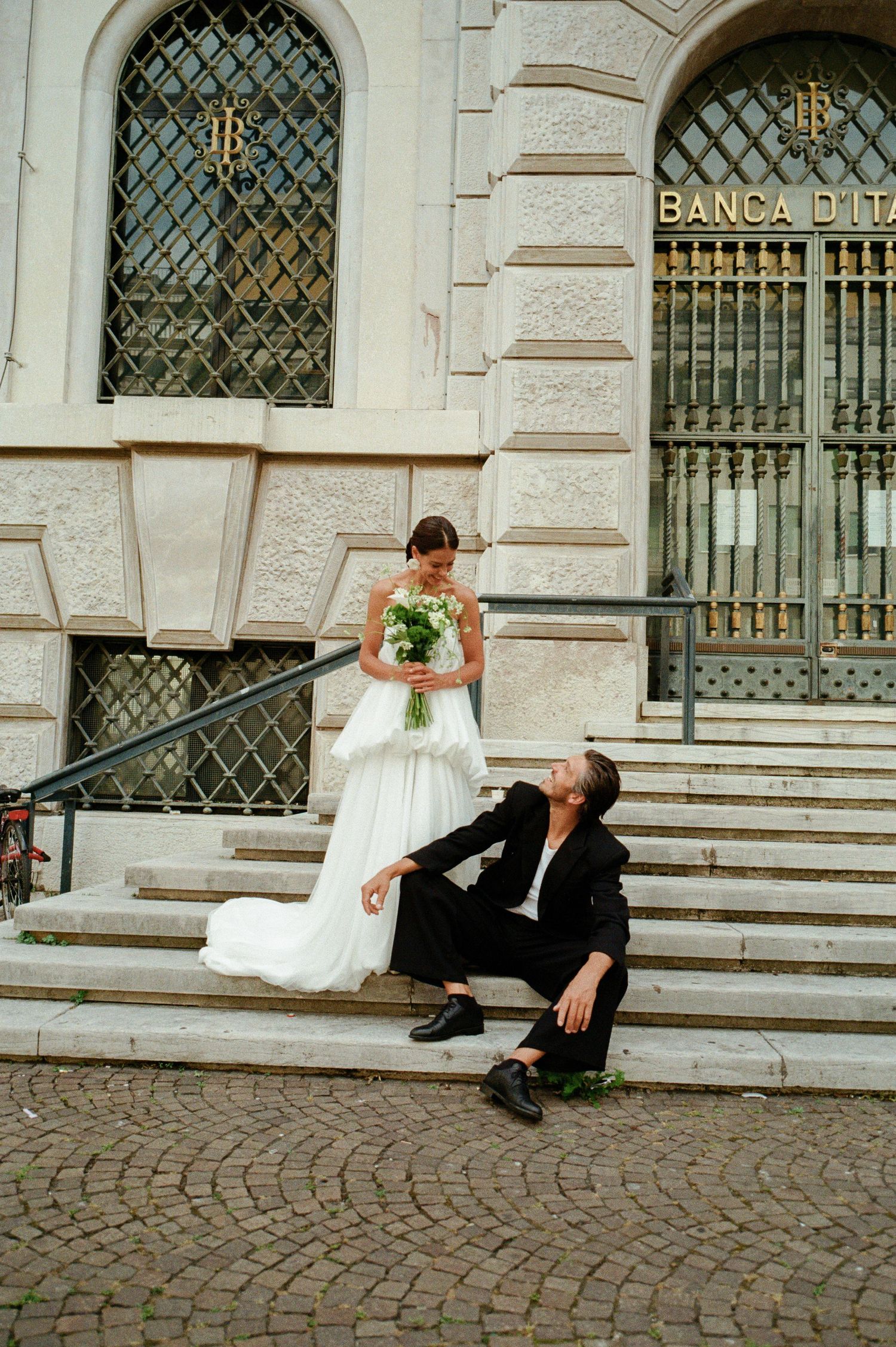 Bride in white gown holds bouquet on steps while groom in black suit sits on cobblestone below her.