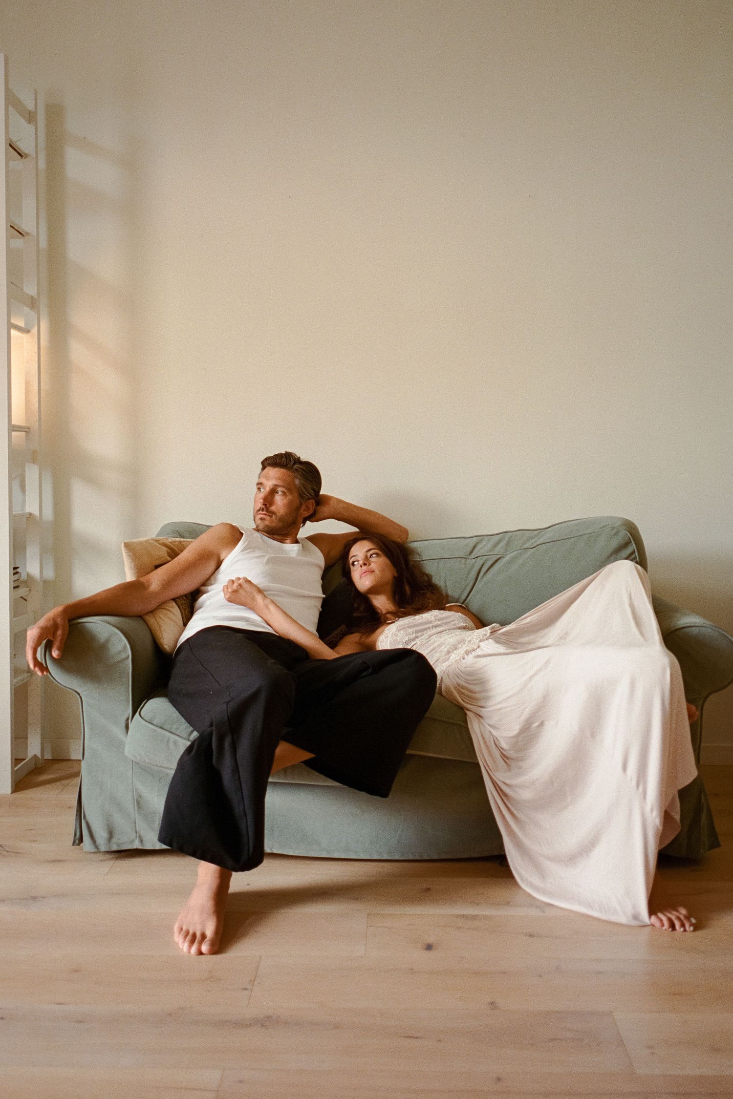 Relaxed couple lounging together on a sage green sofa in a sunlit minimalist room.