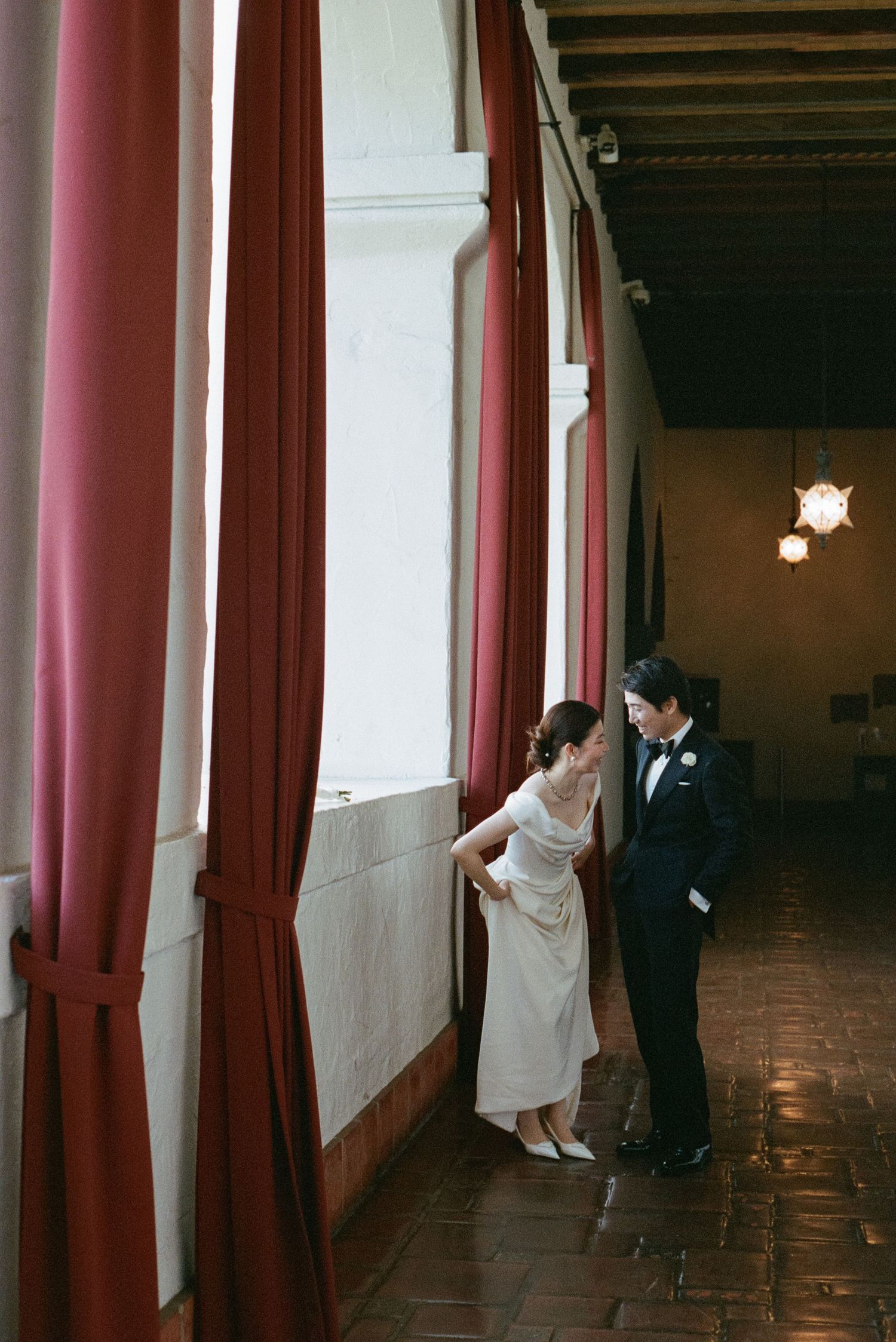 Bride and groom sharing a quiet moment in a hallway with red curtains and warm ambient lighting.