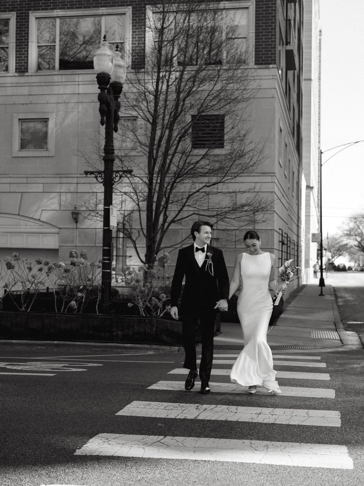 Black and white photo of elegant couple crossing urban street, bride in fitted gown, groom in bow tie tuxedo.