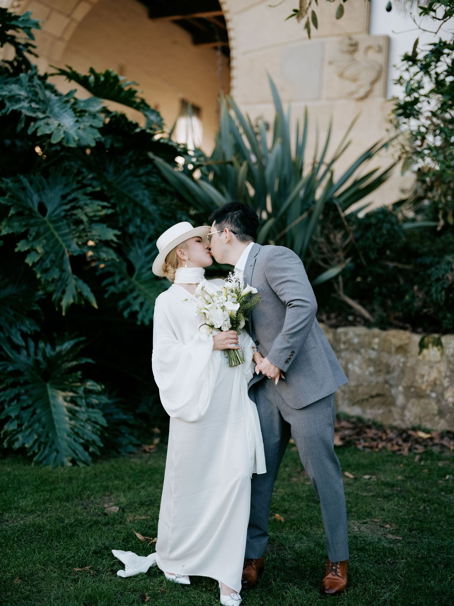 Bride and groom share a kiss on garden lawn, bride in white cape and hat.