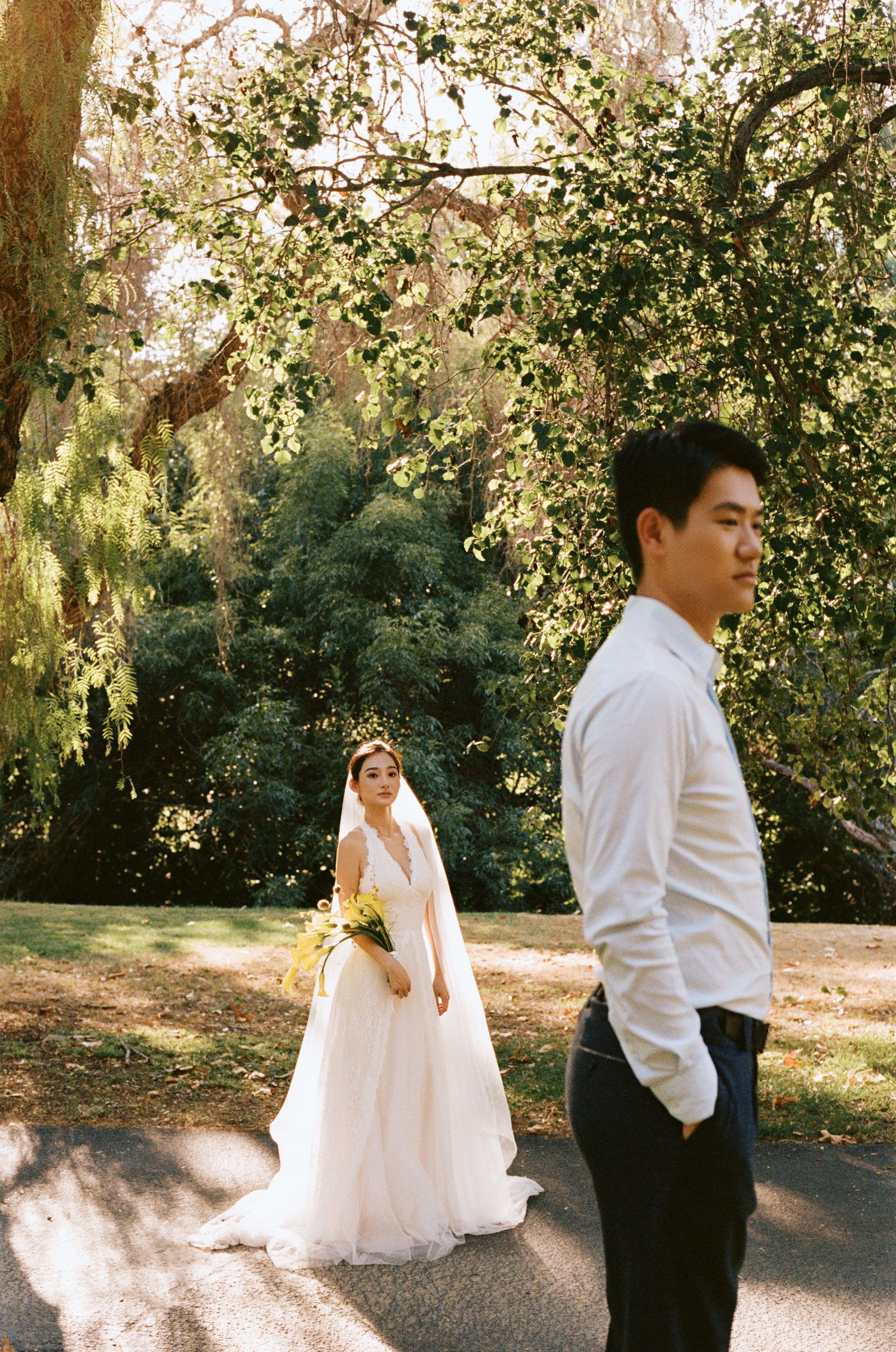 Bride in white gown holds bouquet while groom in white shirt stands in the foreground in a sunlit garden.