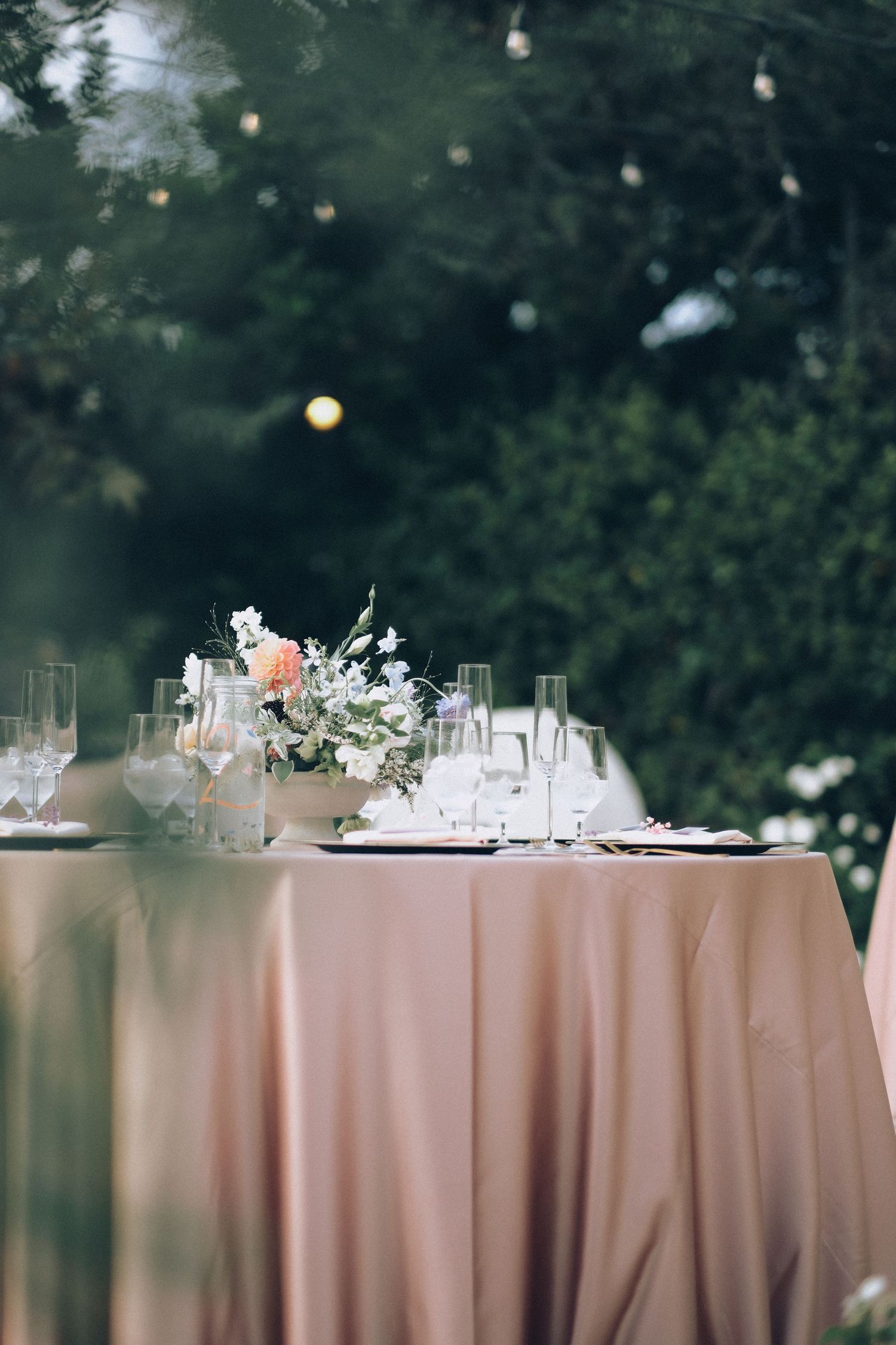 Elegant outdoor wedding reception table with blush pink linen, floral centerpiece, and crystal glassware amid lush greenery.