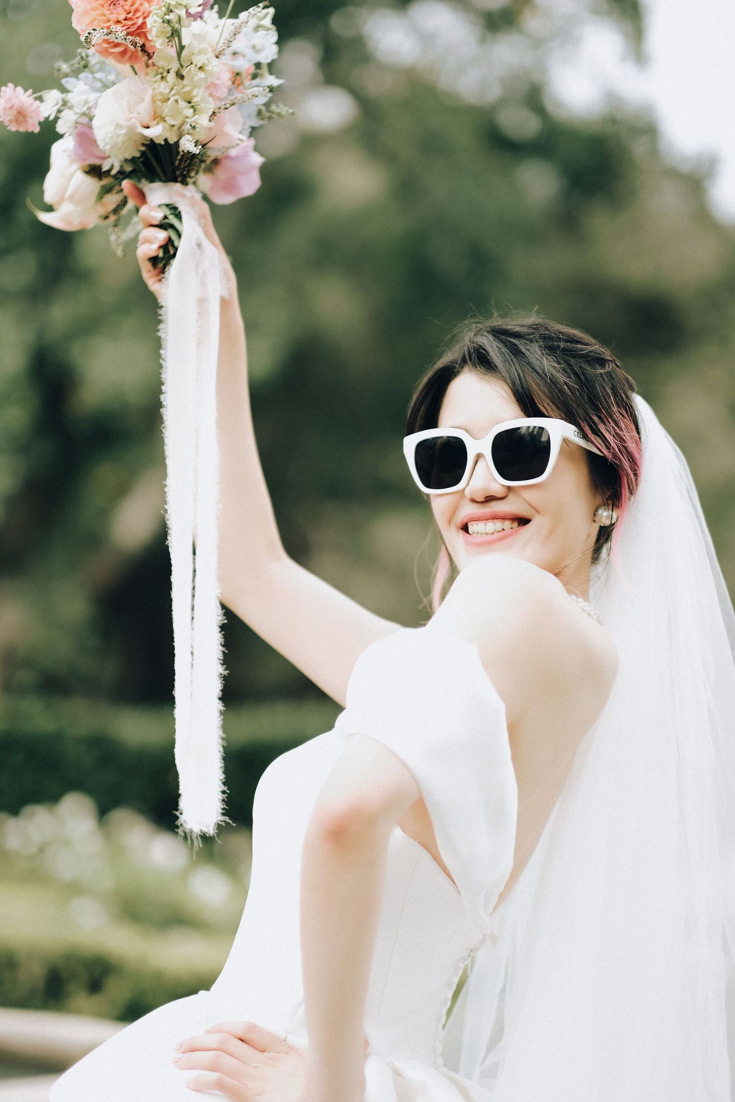 Joyful bride in white sunglasses raises floral bouquet with ribbons outdoors on her wedding day.