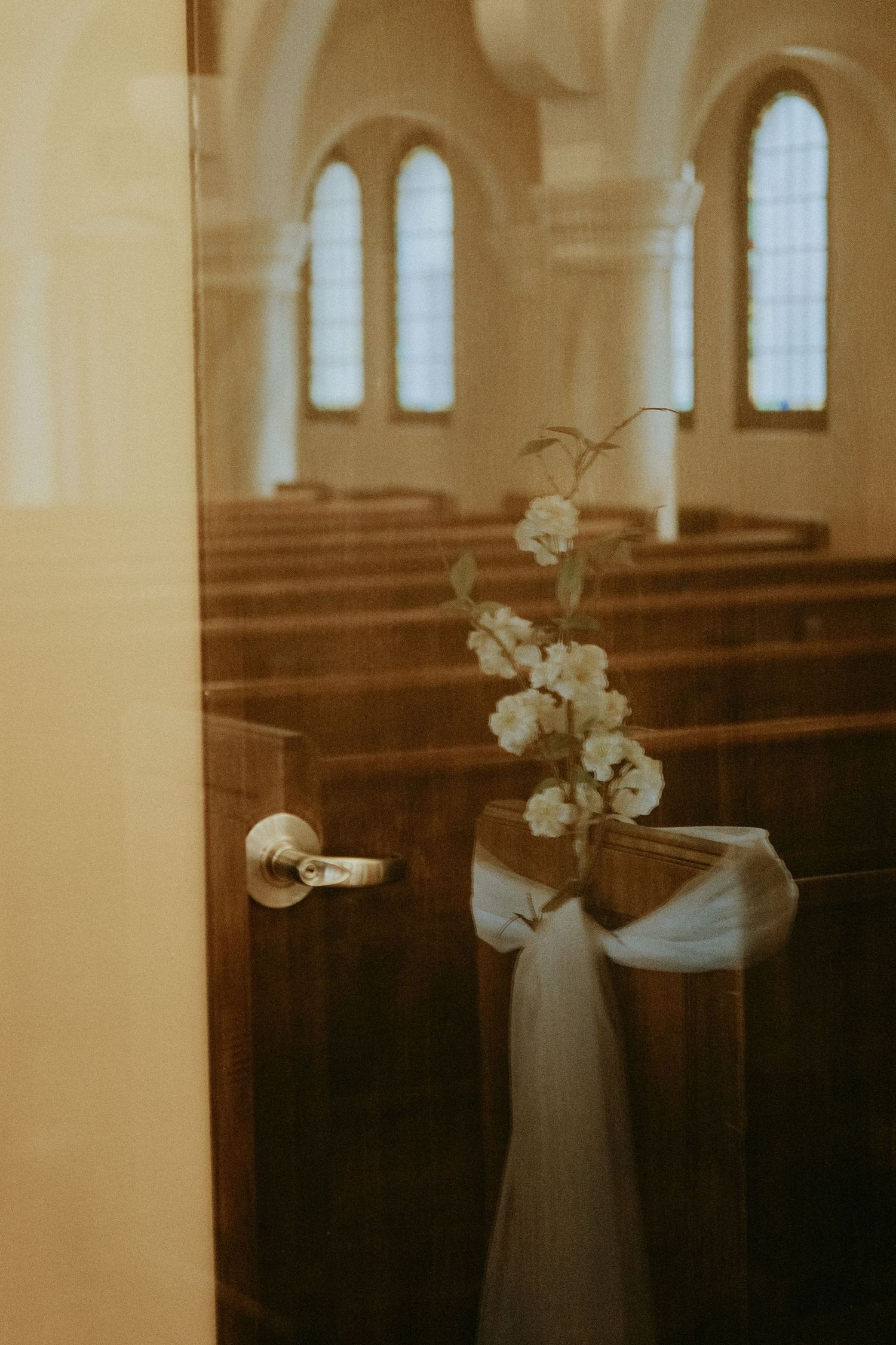 White floral arrangement tied with ribbon on church pew viewed through glass door before wedding ceremony.