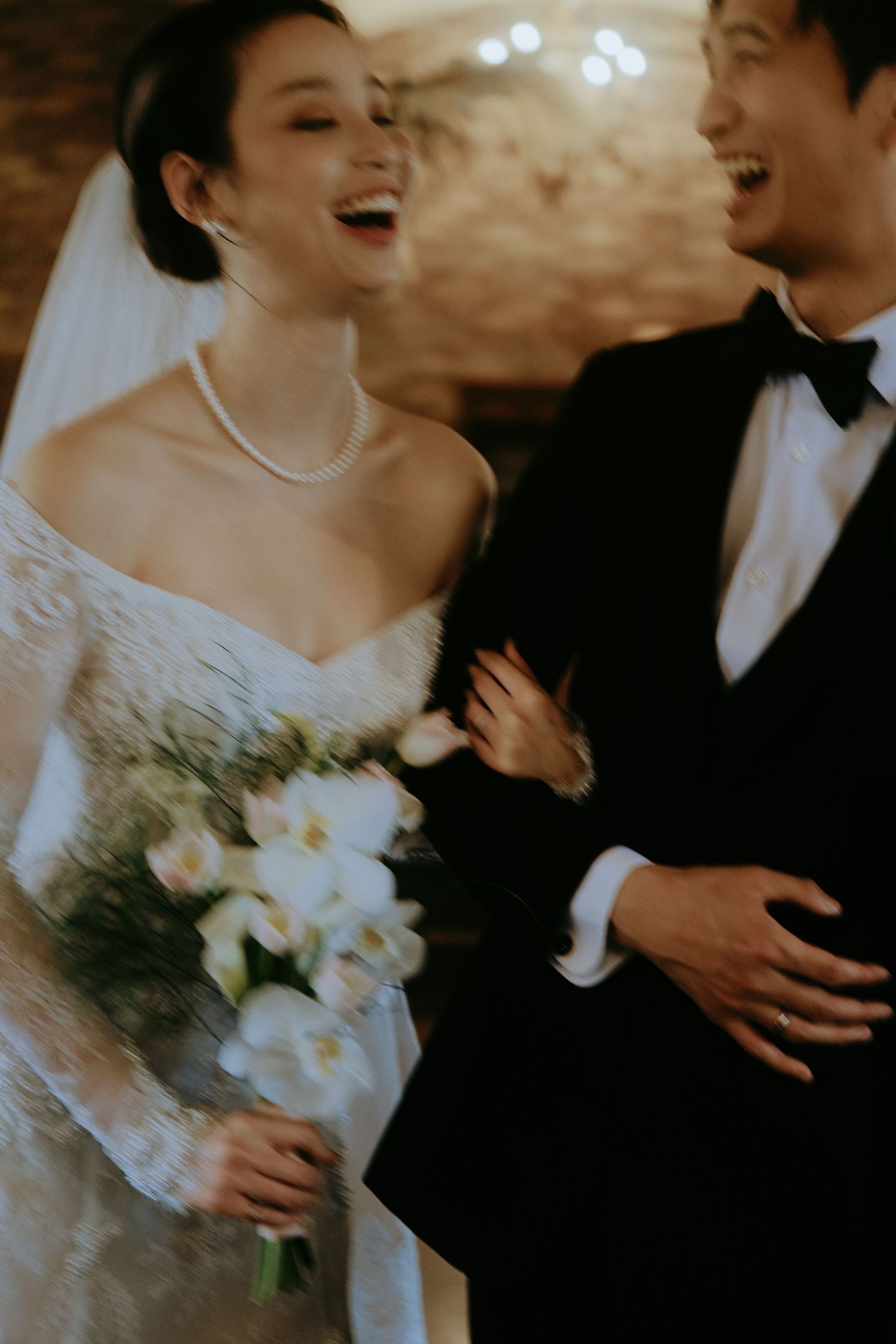 Joyful newlywed Asian couple laughs together, bride holding white floral bouquet in lace wedding gown.
