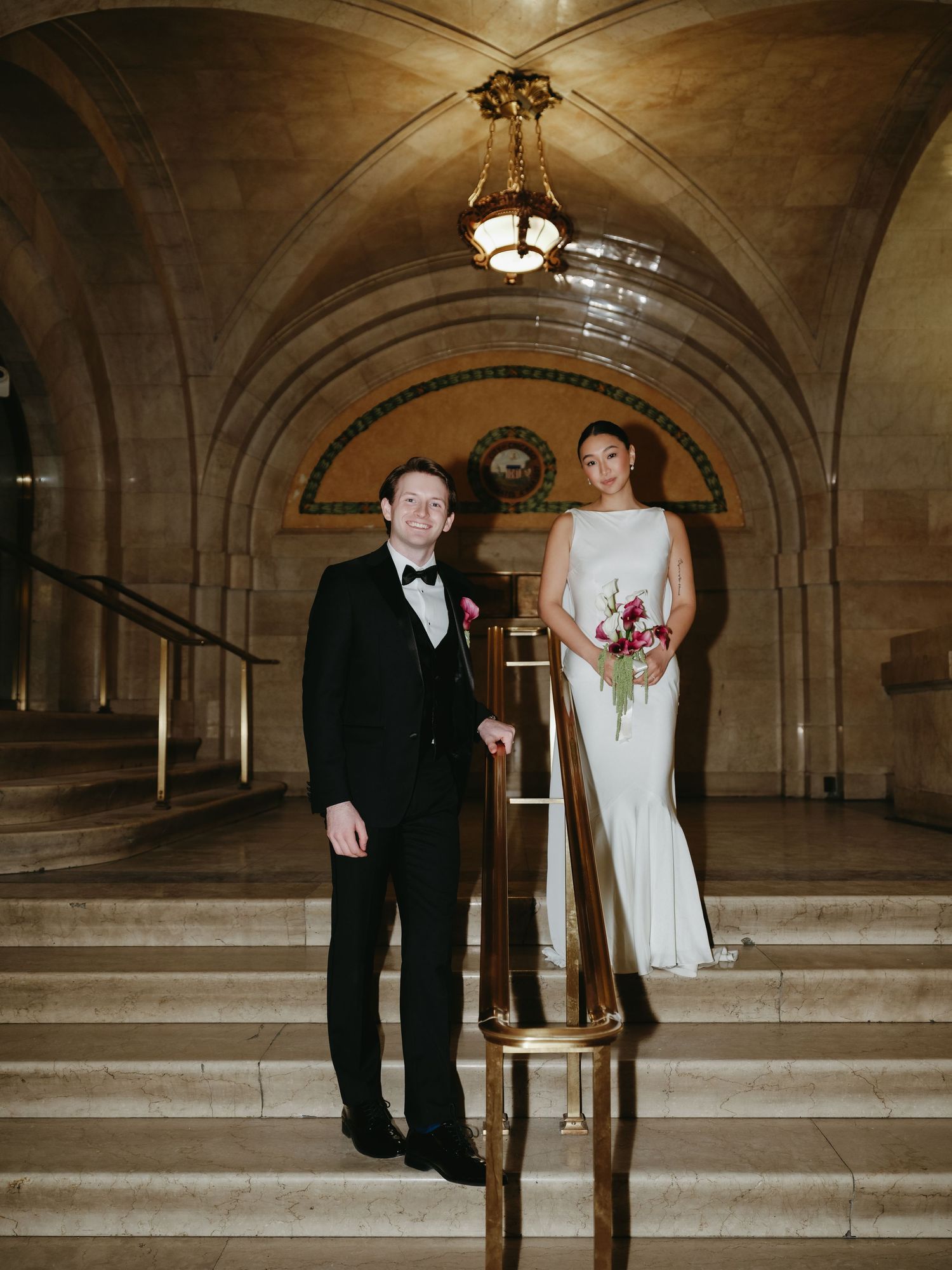 Elegant couple posing on marble stairs inside a grand hall, bride in white gown holding bouquet, groom in black tuxedo.