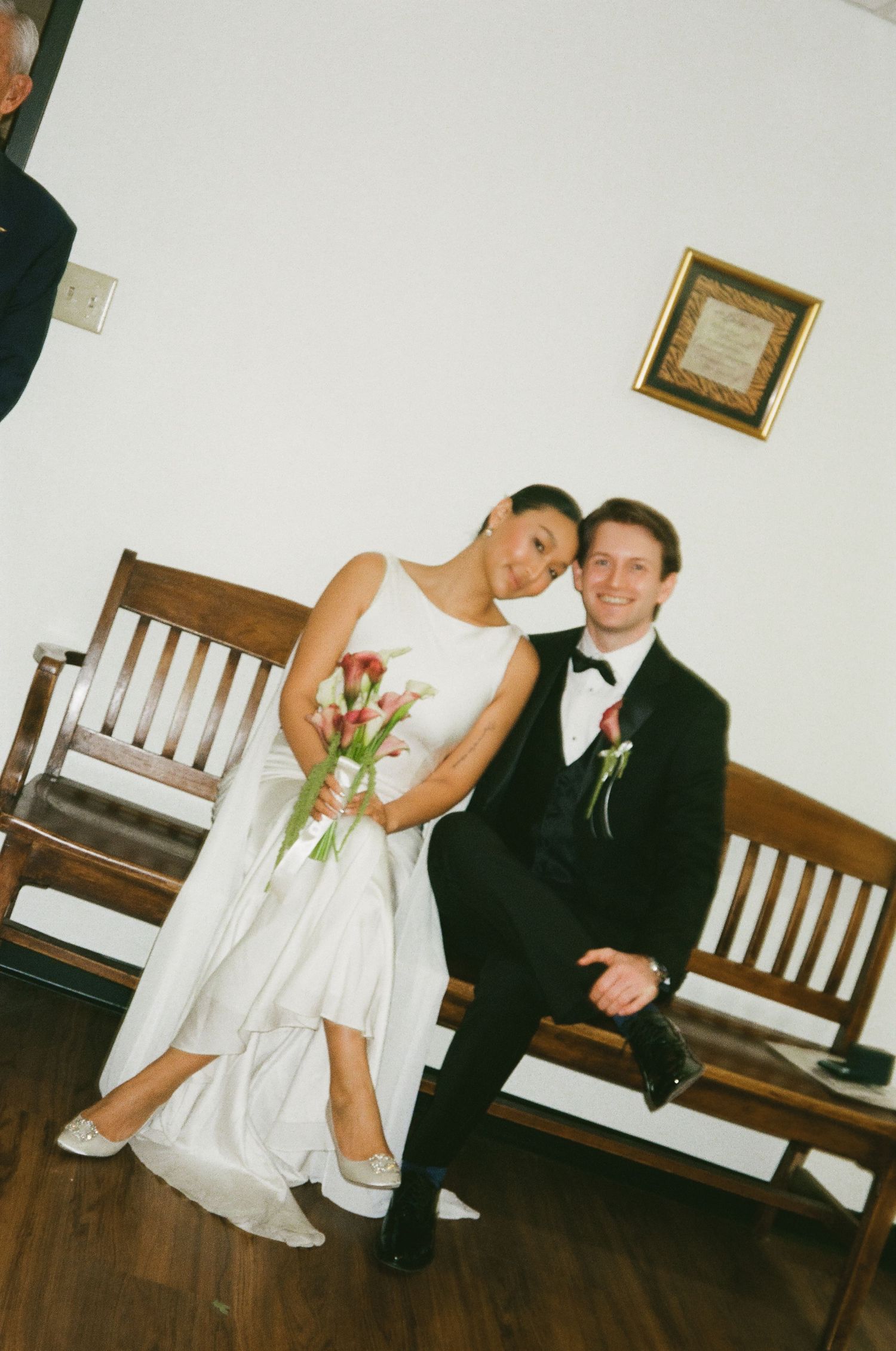 Bride and groom pose on wooden bench, bride holding pink tulip bouquet in a courthouse setting.