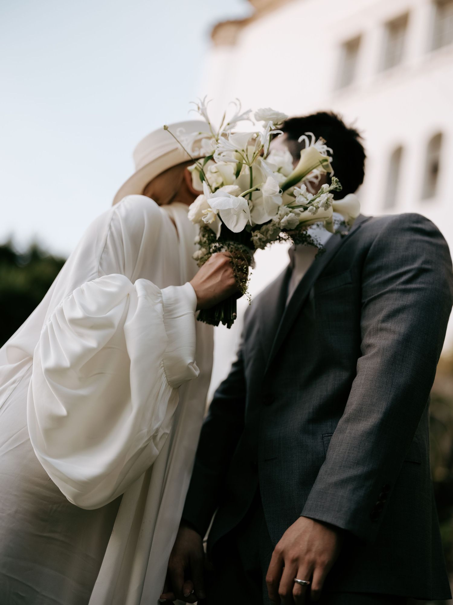 Bride and groom share a kiss hidden behind a lush white floral wedding bouquet.