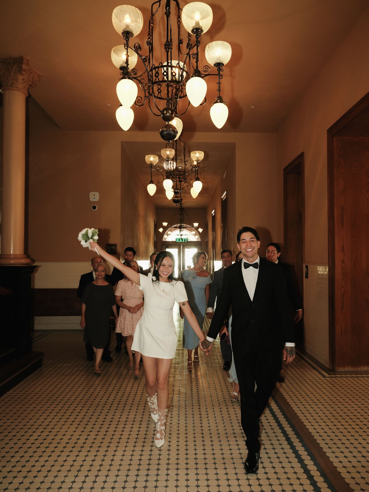 Bride raises bouquet triumphantly while walking with groom through chandelier-lit corridor after ceremony.