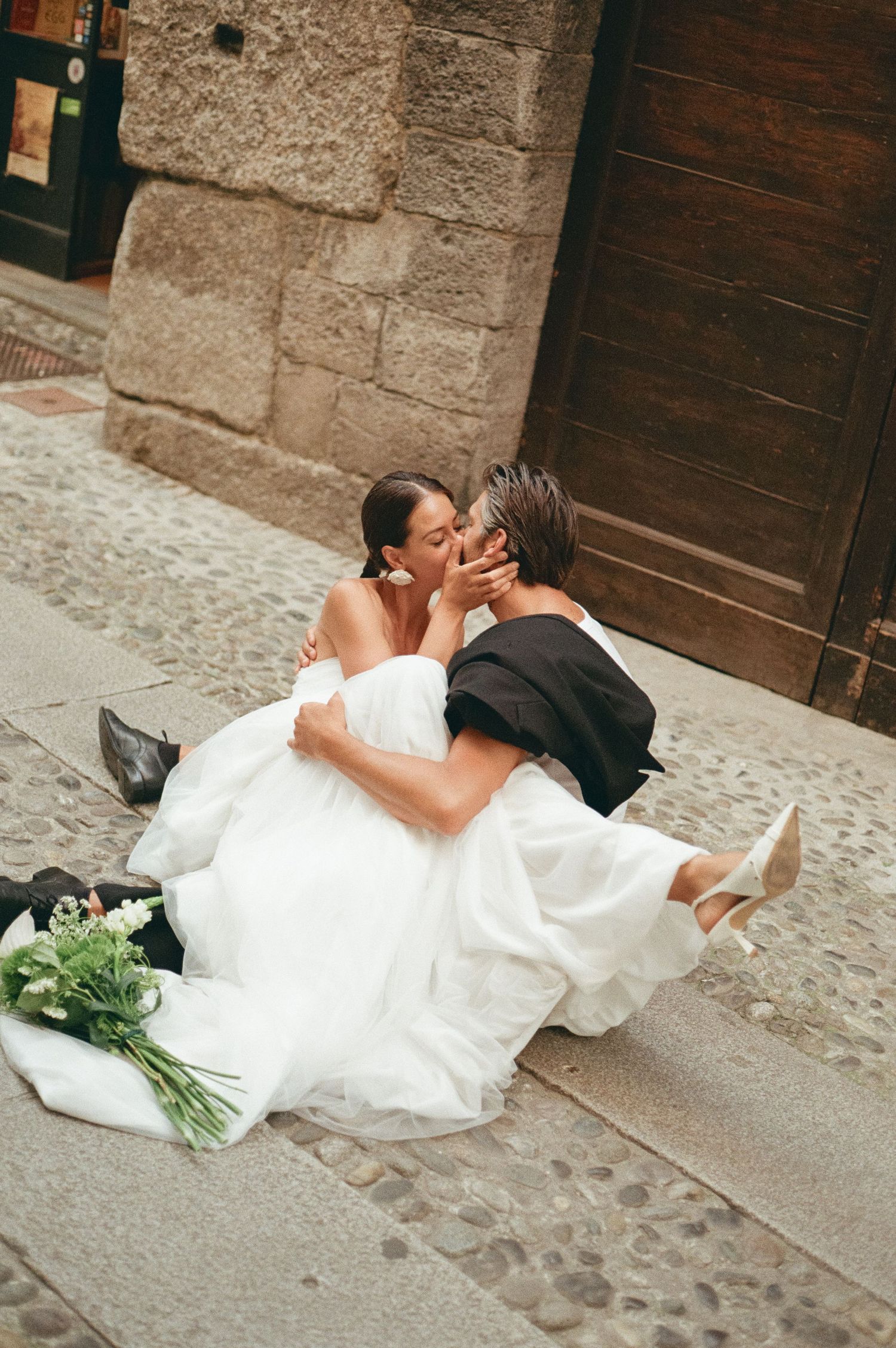Bride and groom sharing a romantic kiss while sitting on cobblestone steps with a bridal bouquet.